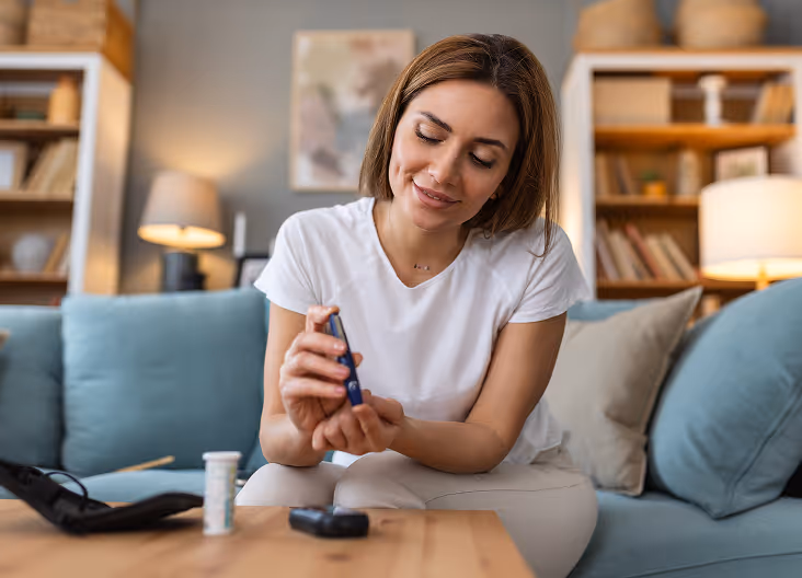 Woman sitting on a couch in a living room, checking her blood sugar with a glucose meter.