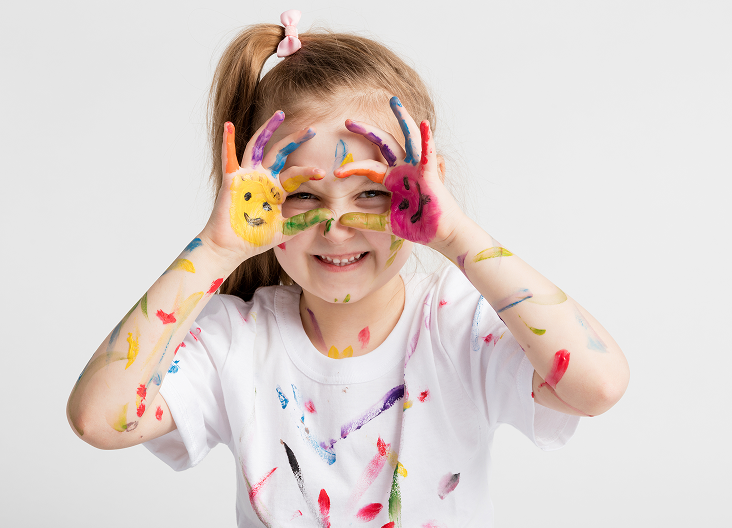Smiling girl with paint-covered hands and arms making circles around her eyes, wearing a white shirt with colorful paint smudges.