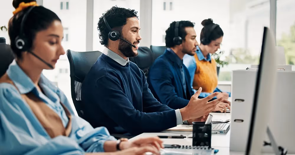 Four customer service representatives wearing headsets working at computers in an office.