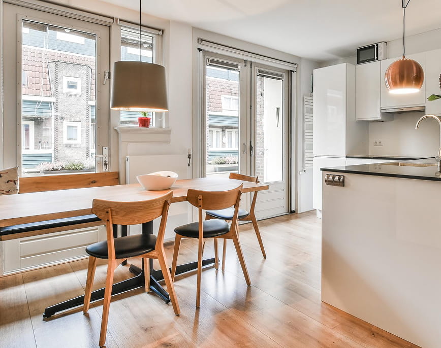 Modern dining area with wooden table, three wooden chairs with black seats, large windows, and a white kitchen with copper pendant lights.