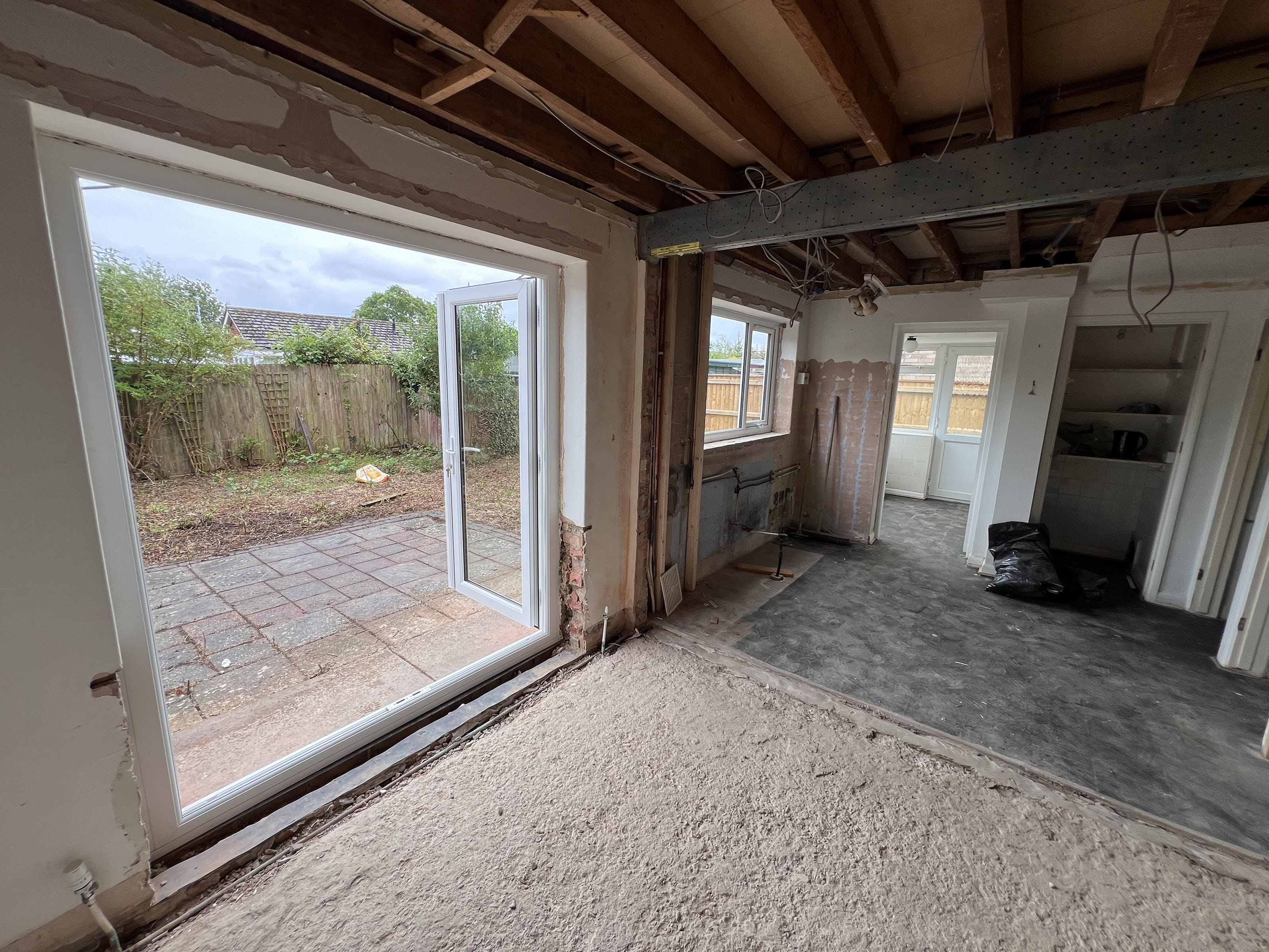 Interior view of a partially renovated room with exposed ceiling beams, open patio doors leading to a backyard, and unfinished floor and walls.