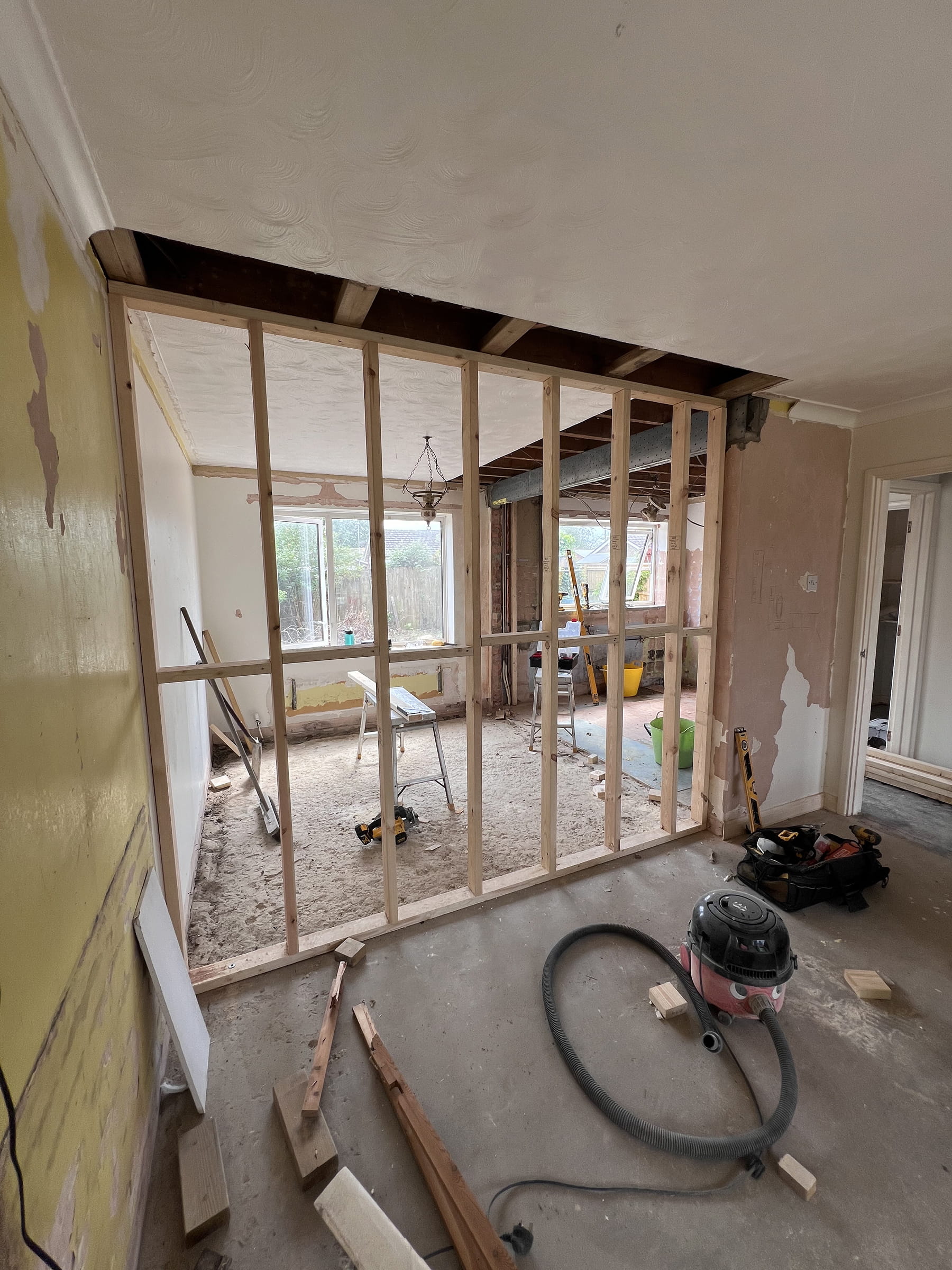 Interior room under renovation showing wooden framing for a new wall, exposed ceiling beams, and construction tools scattered on the floor.