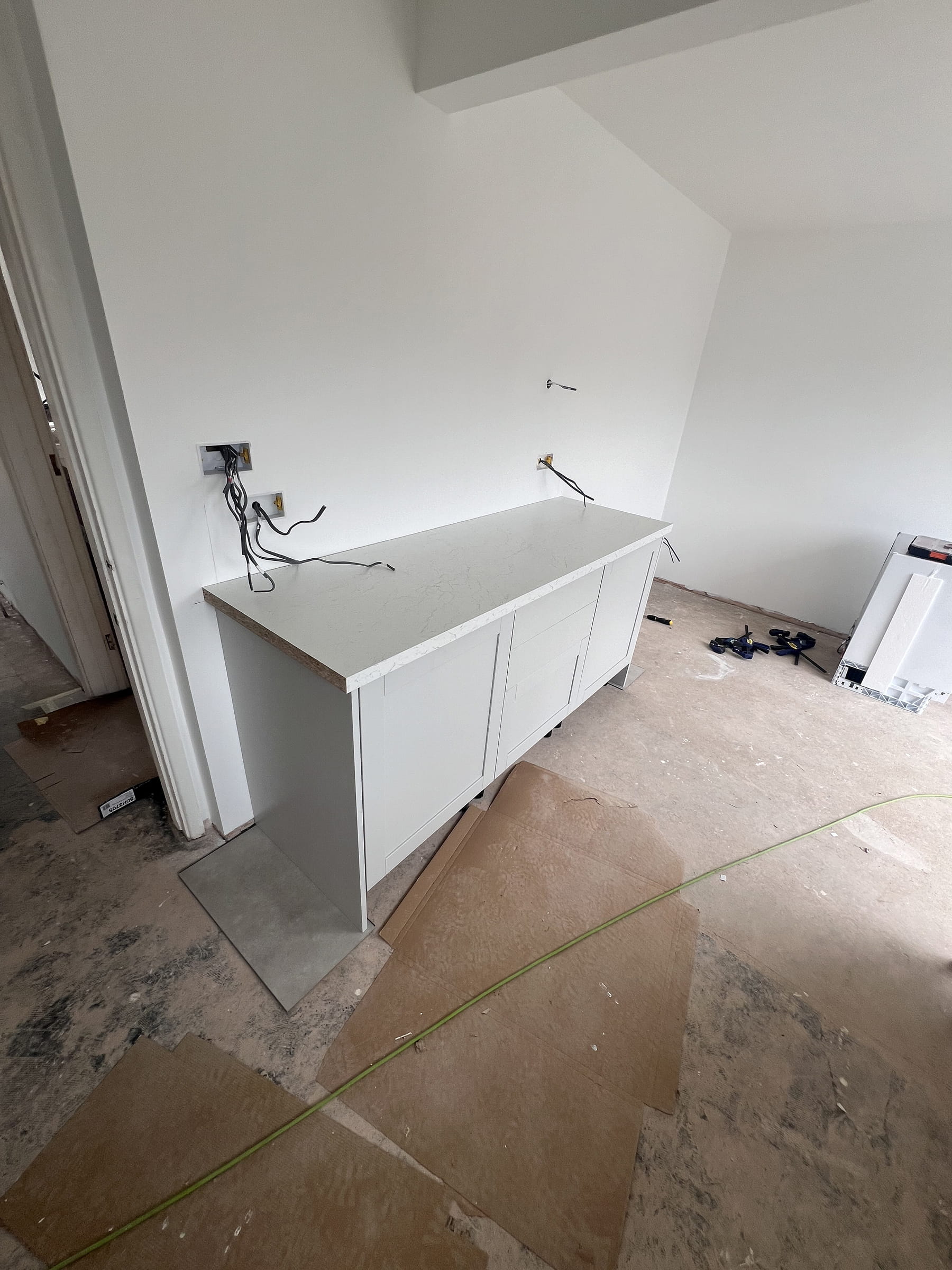 Partially installed white kitchen base cabinet with white marble-effect countertop in an unfinished room with exposed wiring and bare floor.