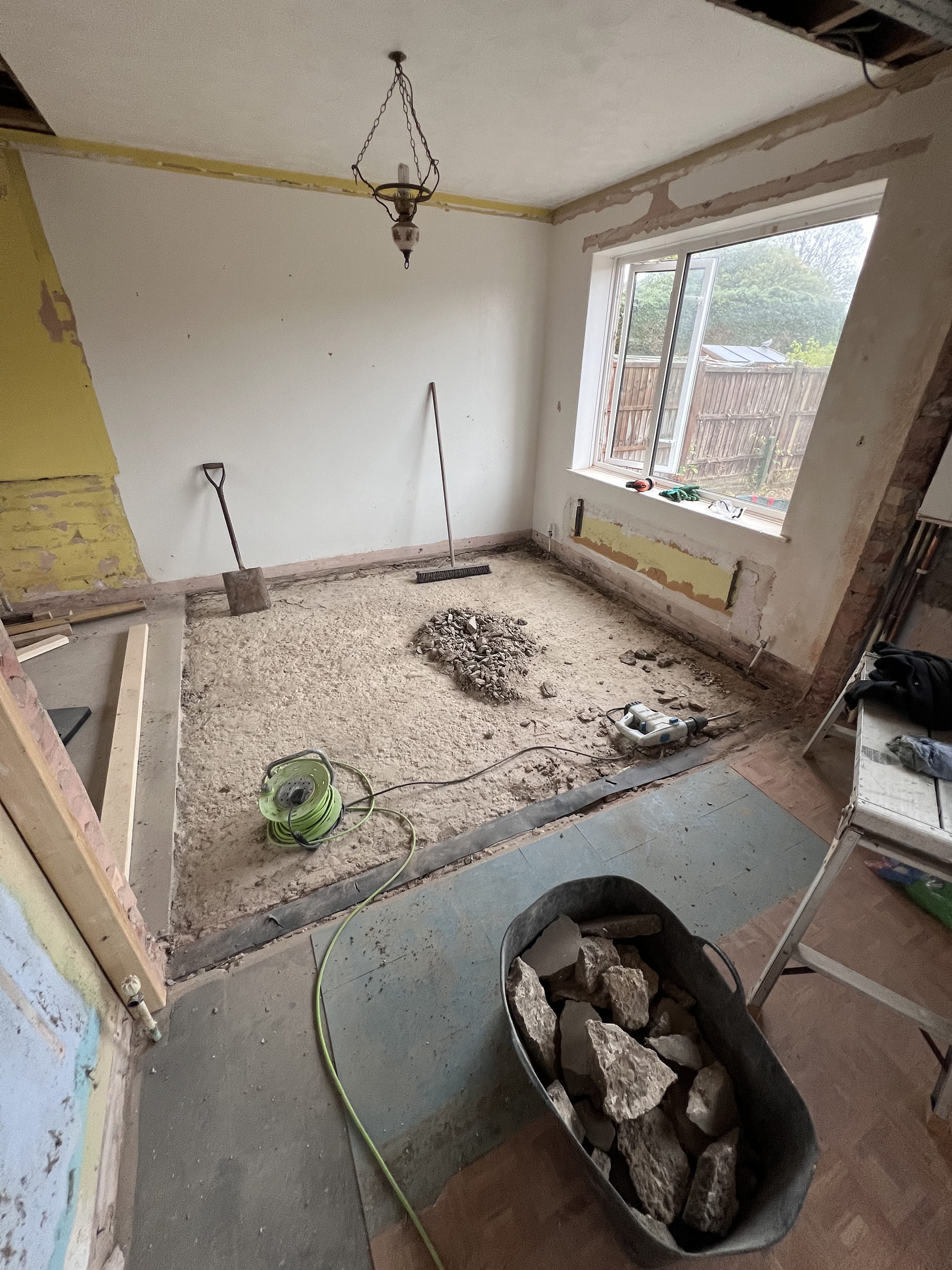 Room under renovation with stripped floor, rubble pile, tools including a shovel, broom, and electric breaker, and a window showing a garden outside.