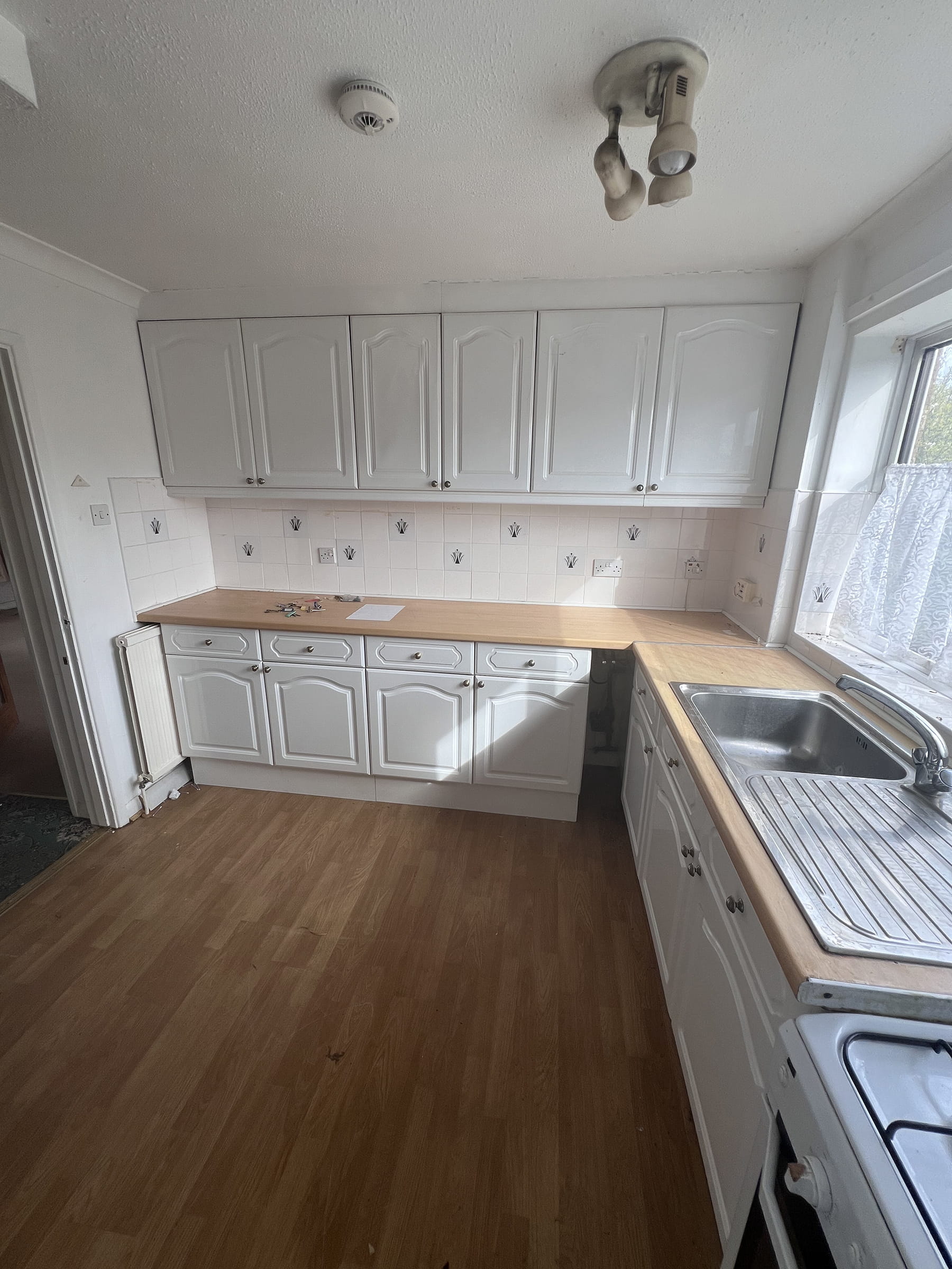 Empty kitchen with white cabinets, wooden countertops, a sink under a window with lace curtains, and a wooden floor.