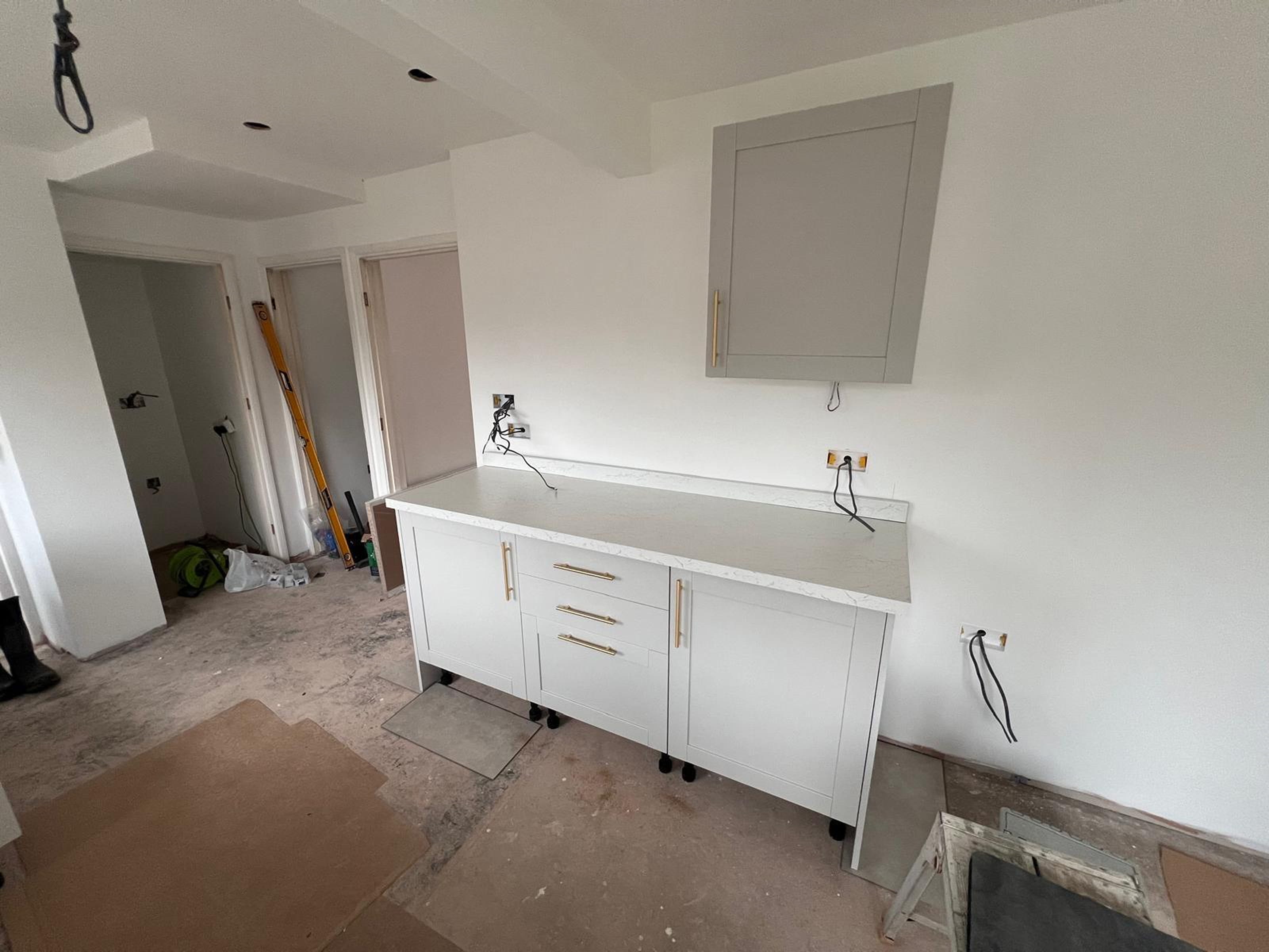 Partially installed white kitchen cabinets with marble countertop in an unfinished room with bare floor and exposed wiring.