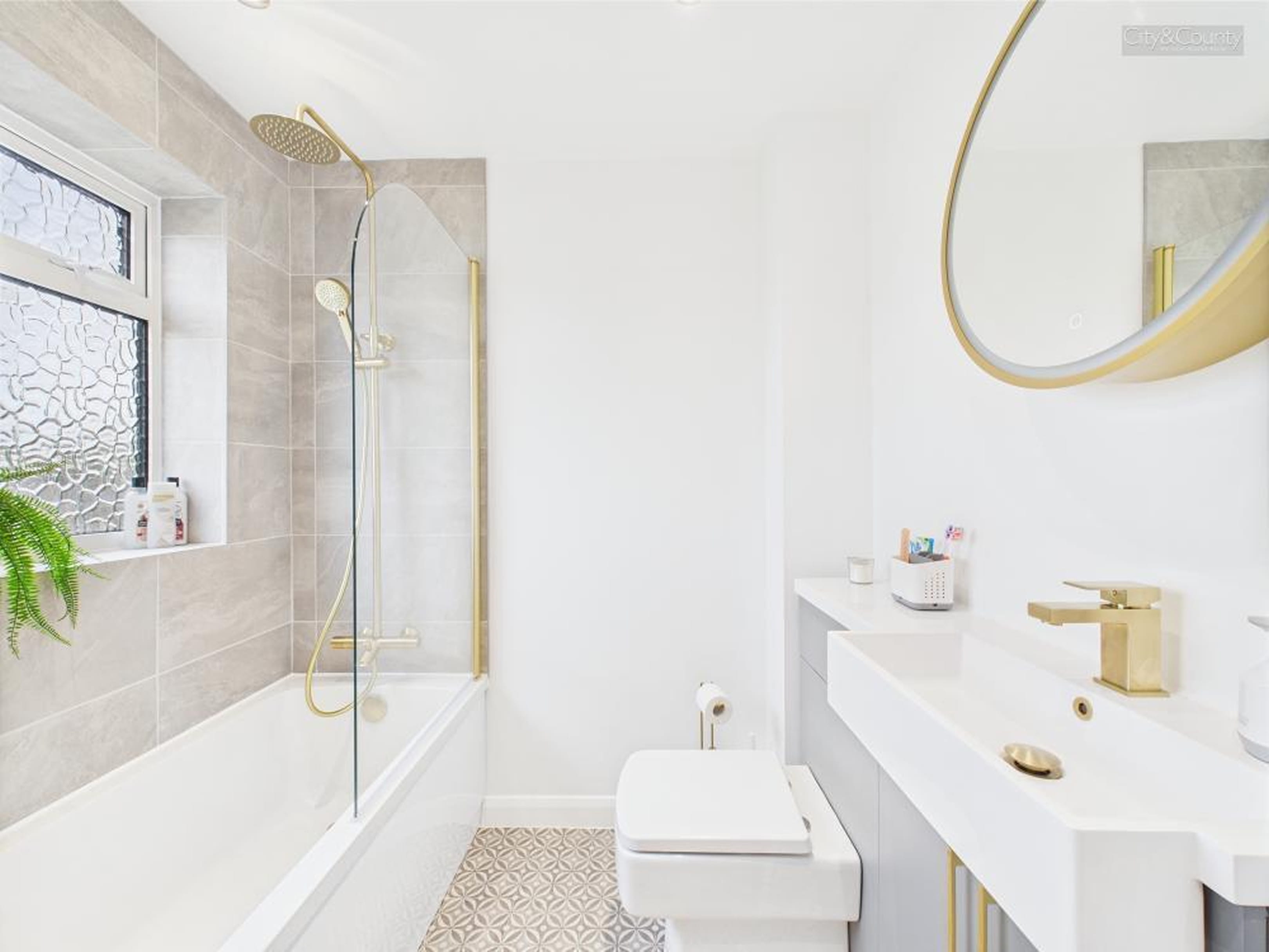 Modern bathroom with white bathtub, gold shower fixtures, patterned floor tiles, white toilet, and a white sink with gold faucet under a round mirror.