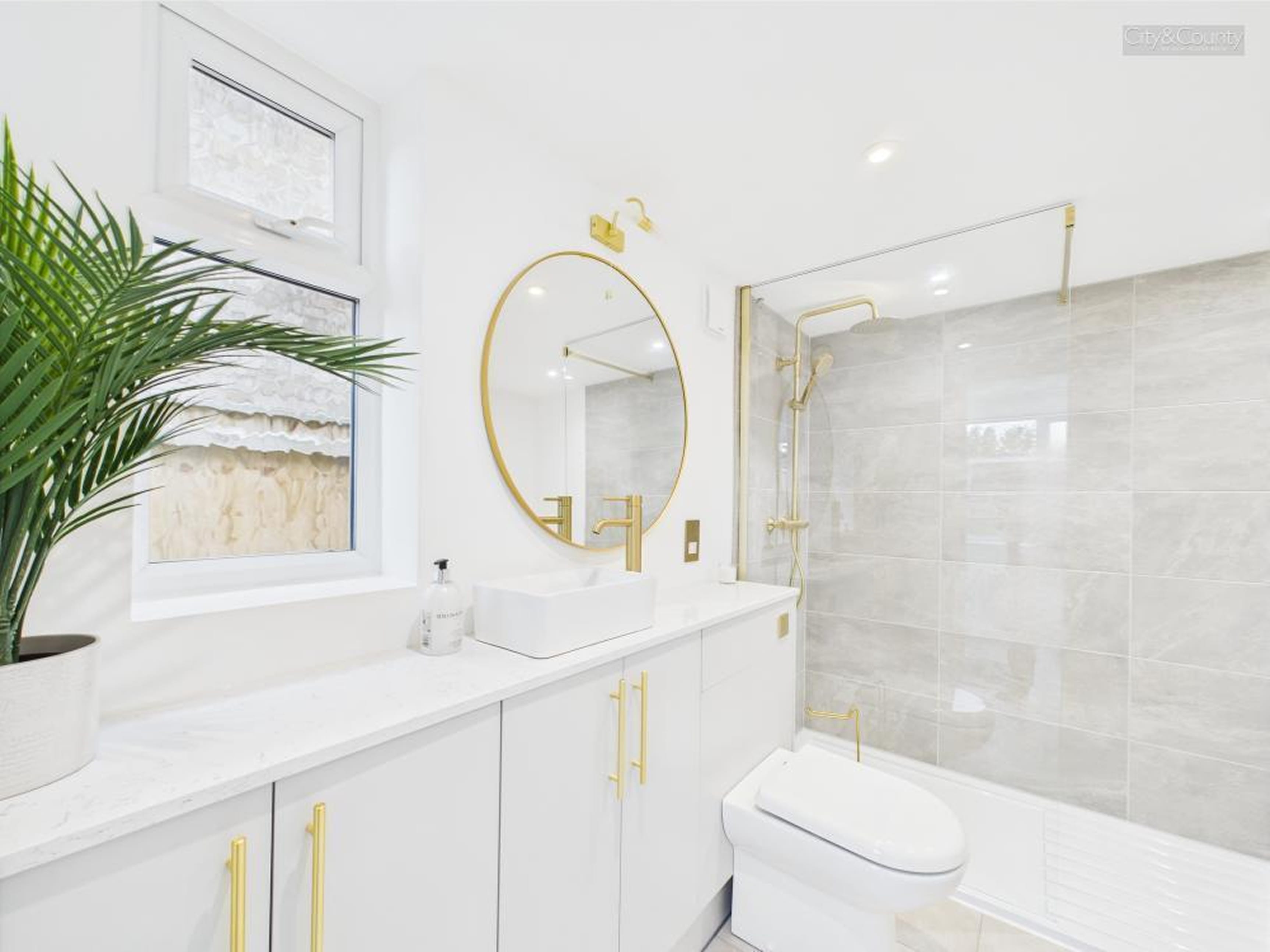 Modern bathroom with white cabinet, gold fixtures, round mirror, potted plant by frosted window, and shower with glass partition and grey tiles.