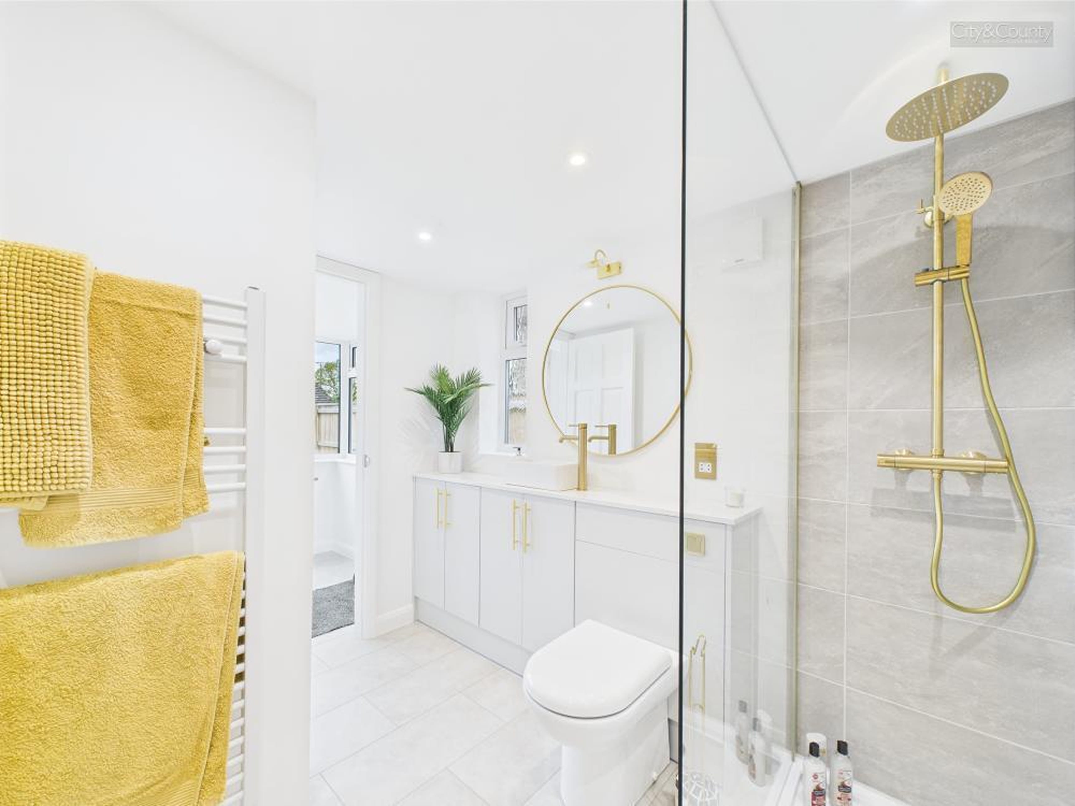 Bright modern bathroom with yellow towels on heated rail, white vanity with gold handles, round mirror, plant, and gold shower fixtures.