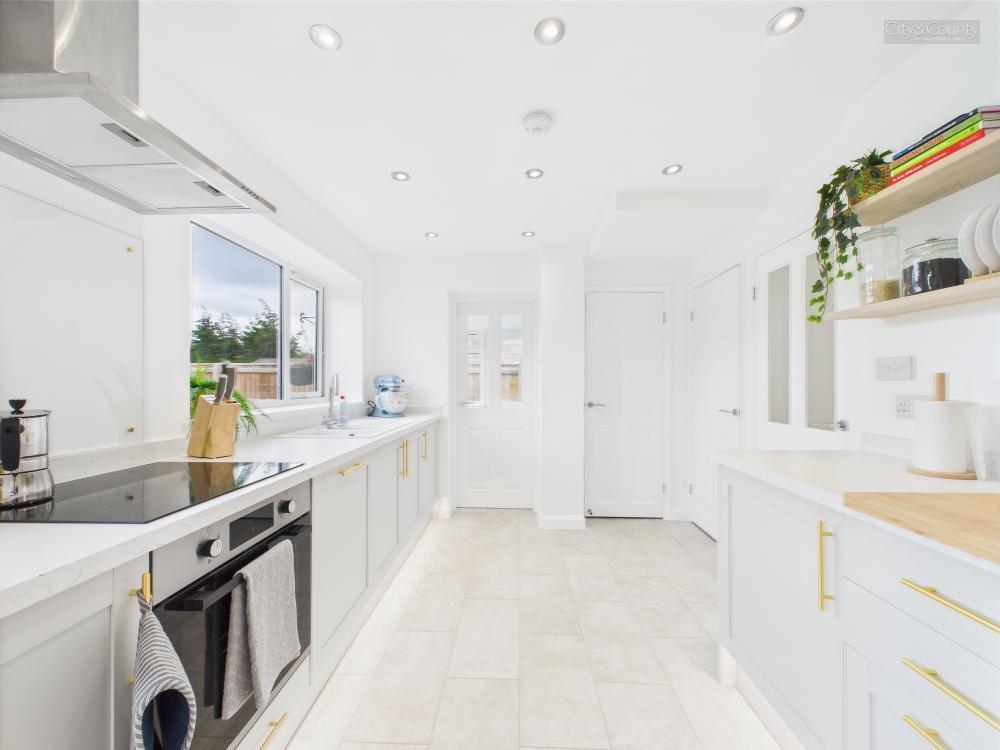 Bright modern kitchen with white walls, grey cabinets with gold handles, built-in oven, induction stove, large window, and open shelves with plants and jars.