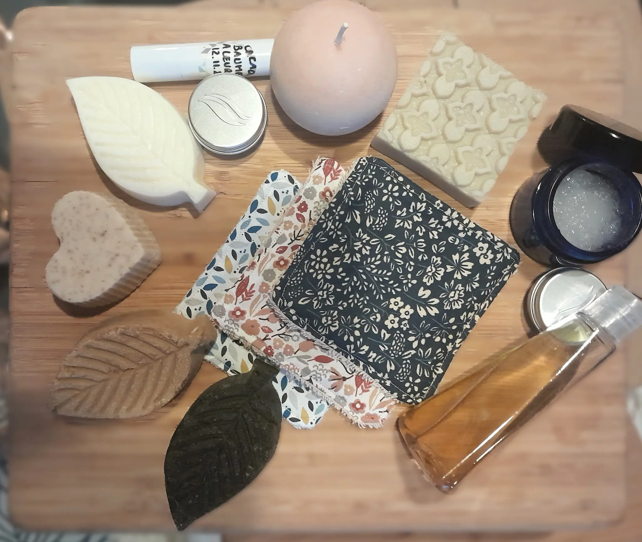 Collection of handmade soap bars shaped like leaves and hearts, floral-patterned fabric squares, a round candle, small containers, and a bottle of liquid on a wooden surface.