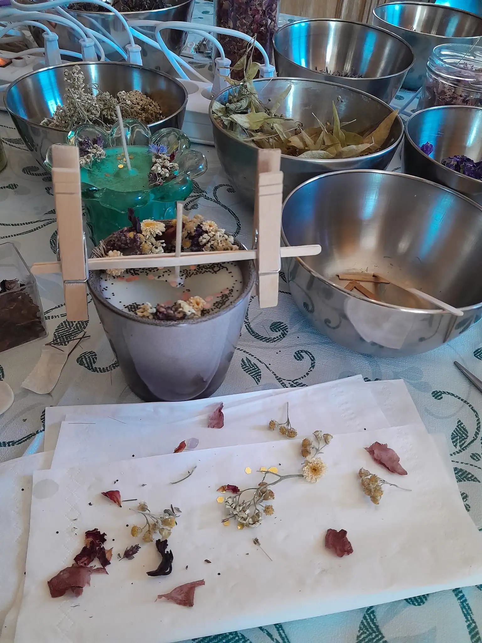 Candle-making setup with dried flowers, metal bowls, and two candles in progress, one held by clothespins on a stick.
