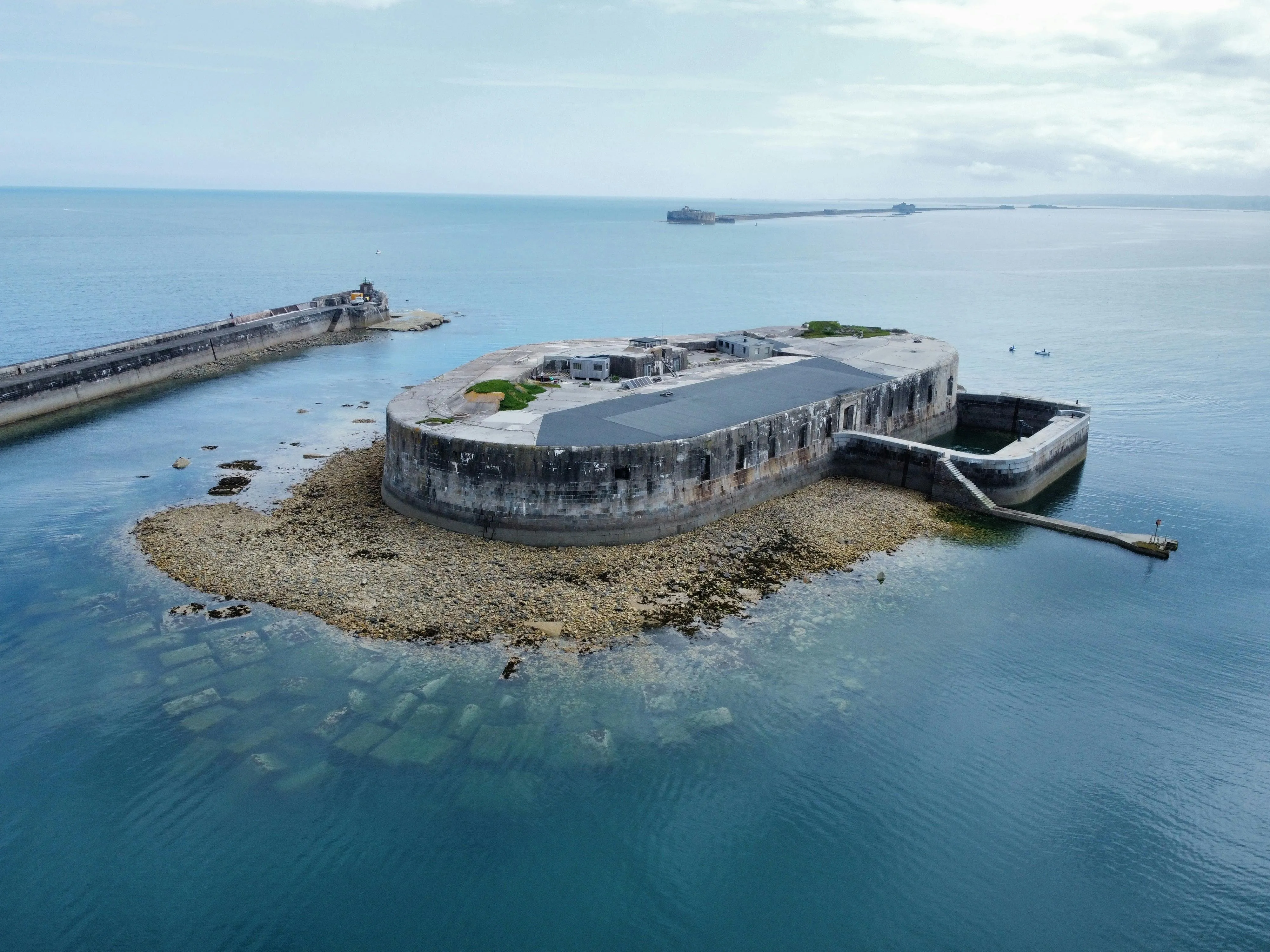 Stone fort on a rocky islet surrounded by calm sea with a breakwater and distant shore under a cloudy sky.