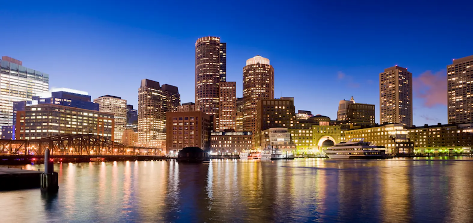Illuminated city skyline at dusk with reflections on calm waterfront.