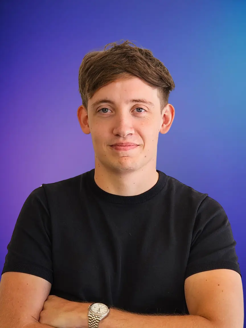 Jonge man met kastanjebruin haar en een zwarte T-shirt, armen gekruist, voor een blauwe achtergrond.