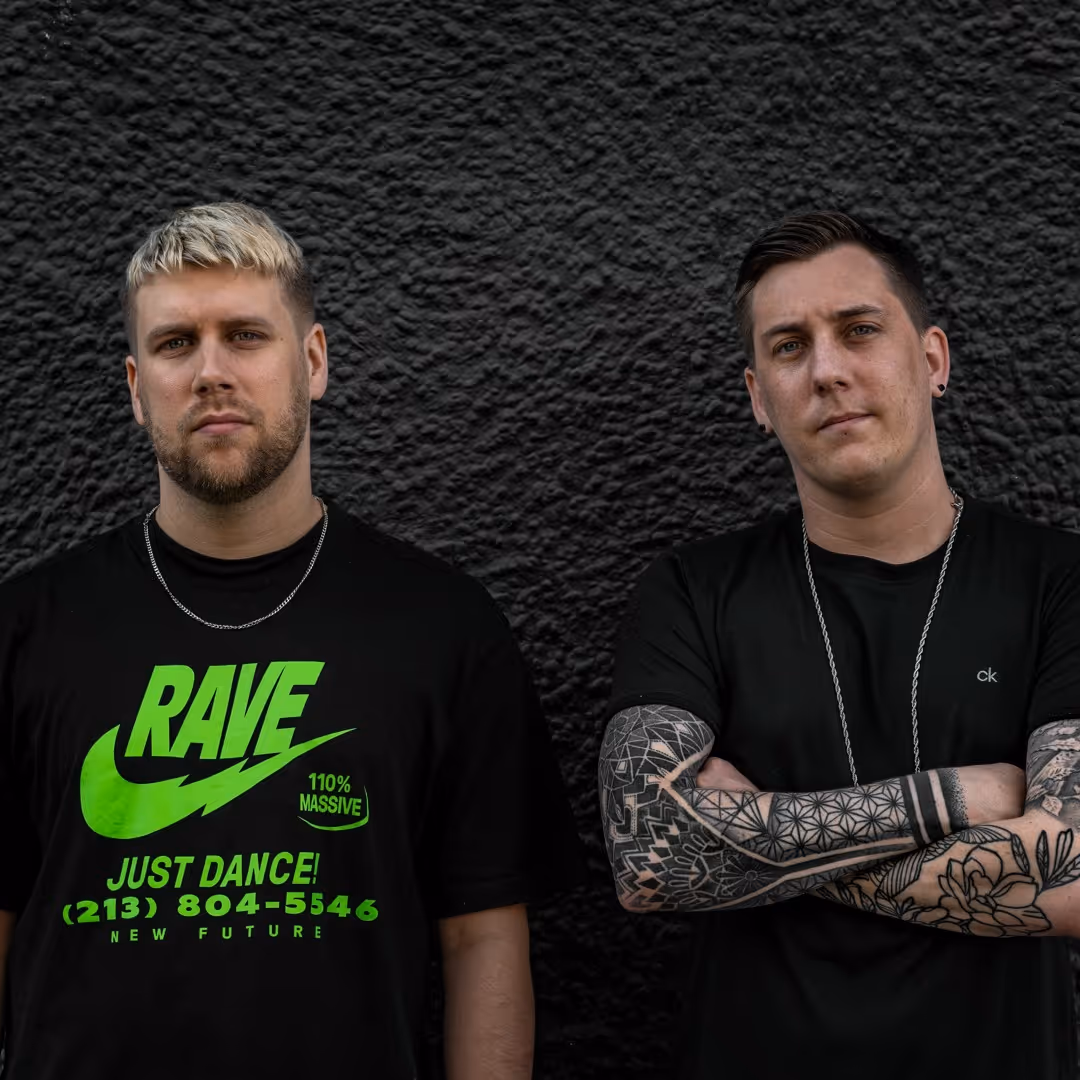 Two men standing against a black textured wall; left man wears a black t-shirt with neon green text and has short blond hair, right man has tattooed arms crossed and short dark hair.