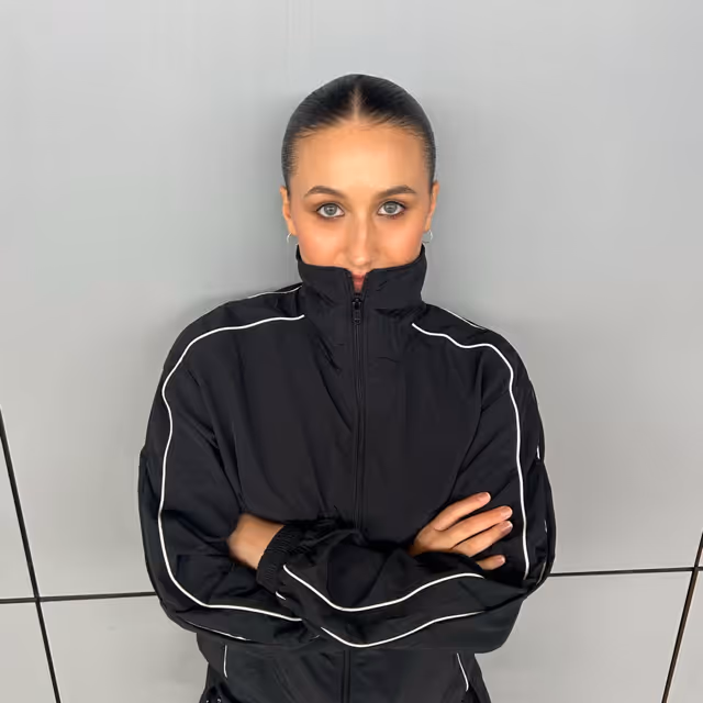 Woman with slicked-back hair wearing a black zip-up jacket with white piping, standing with arms crossed against a light gray tiled wall.