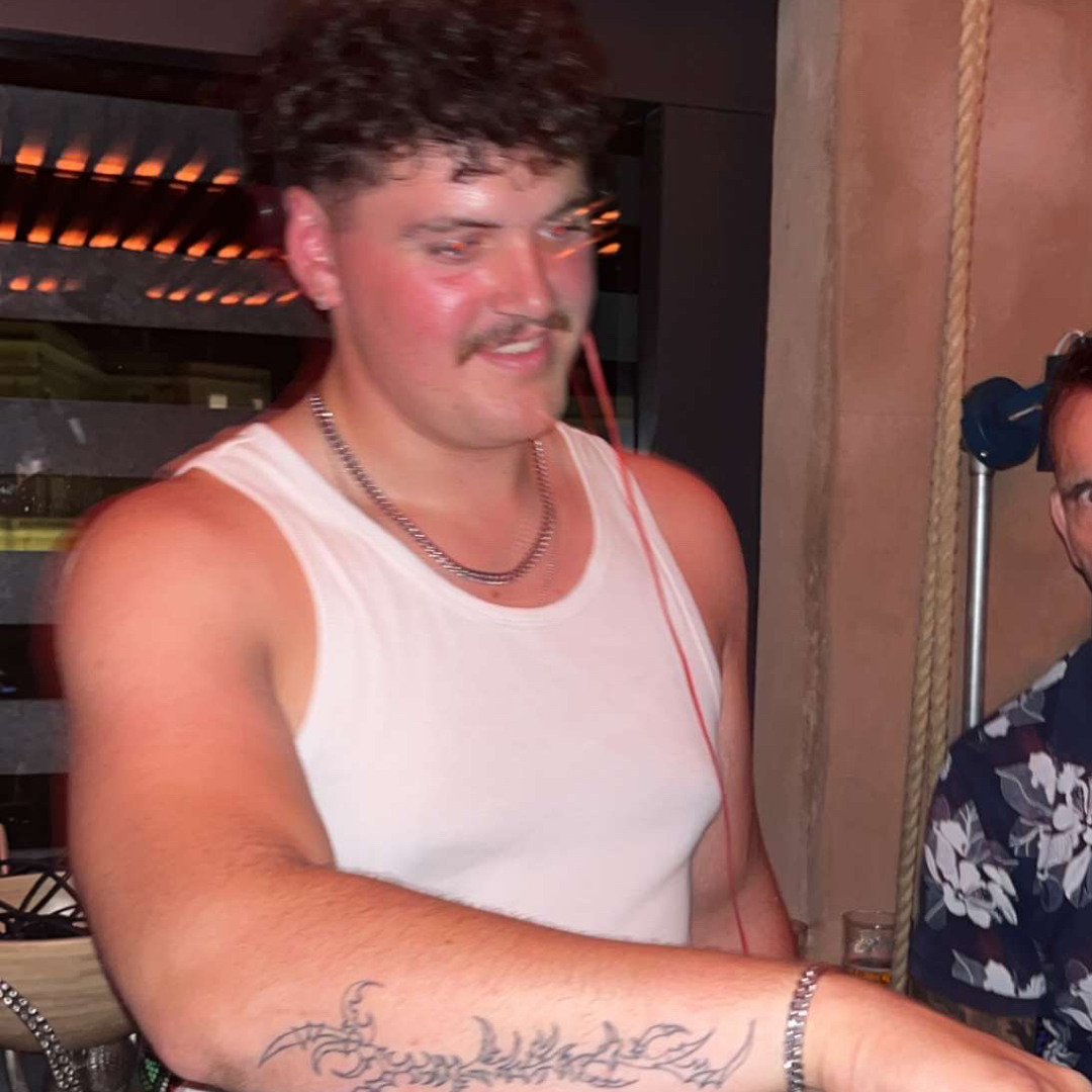 Man in a white tank top with a chain necklace and a tattoo on his left forearm.