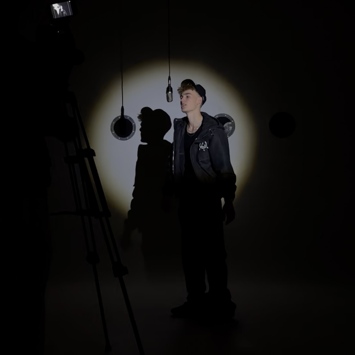 Young man wearing a baseball cap and dark jacket standing in a spotlight in front of hanging microphones, with a camera recording him on the left.