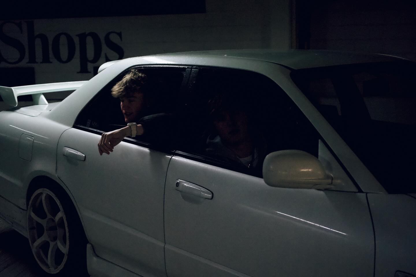 Two young men sitting inside a parked white car at night, one with his arm resting out the open window.