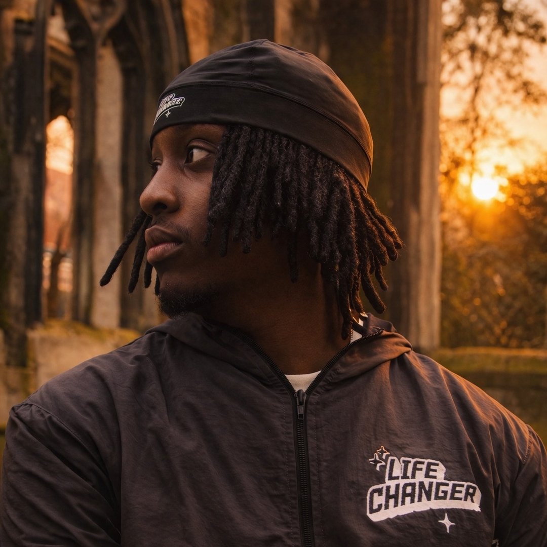 Young man with dreadlocks wearing a black headband and jacket with 'Life Changer' text looks to the side at sunset near stone arches.