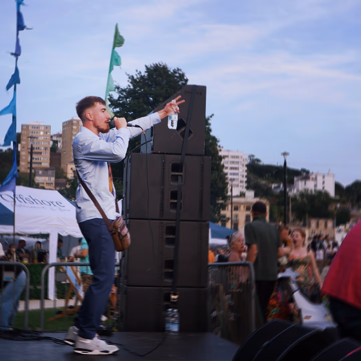 Man with a microphone and water bottle performing on an outdoor stage at dusk with a crowd and city buildings in the background.