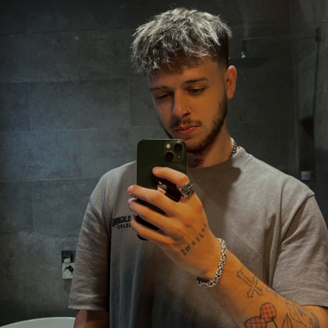 Young man with two-tone hair taking a selfie in a bathroom mirror, wearing a gray shirt and displaying tattoos on his arm and hand.