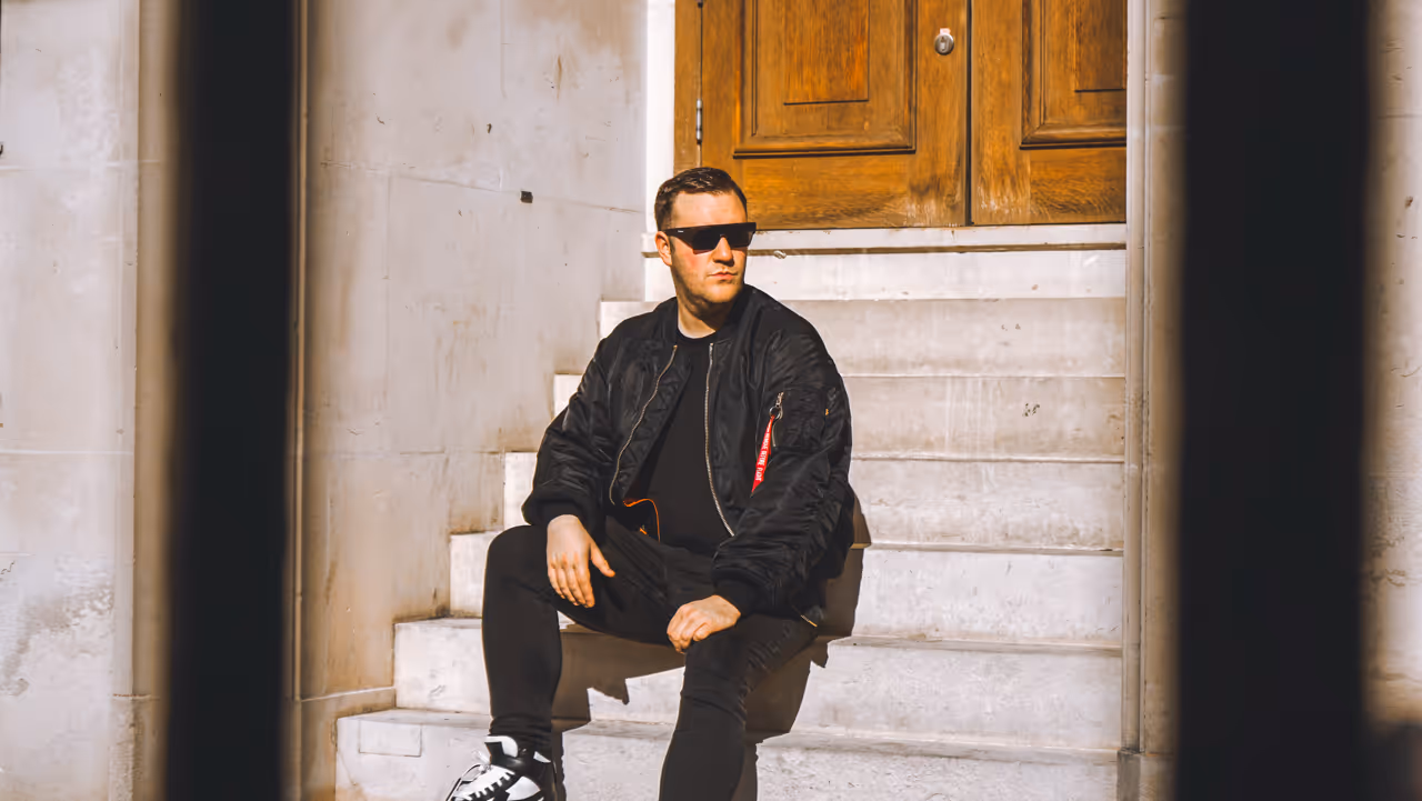 Man wearing black jacket, sunglasses, and sneakers sitting on white stone steps in front of a wooden door.