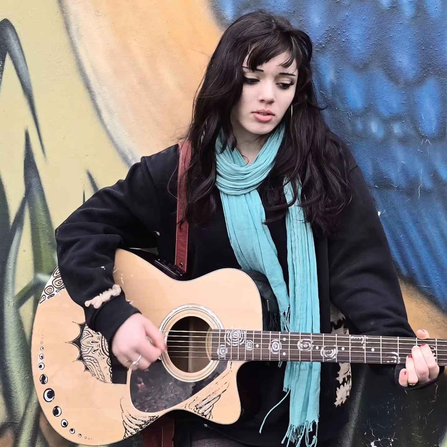 Young woman with dark hair wearing a black sweater and turquoise scarf playing a decorated acoustic guitar against a colorful graffiti wall.