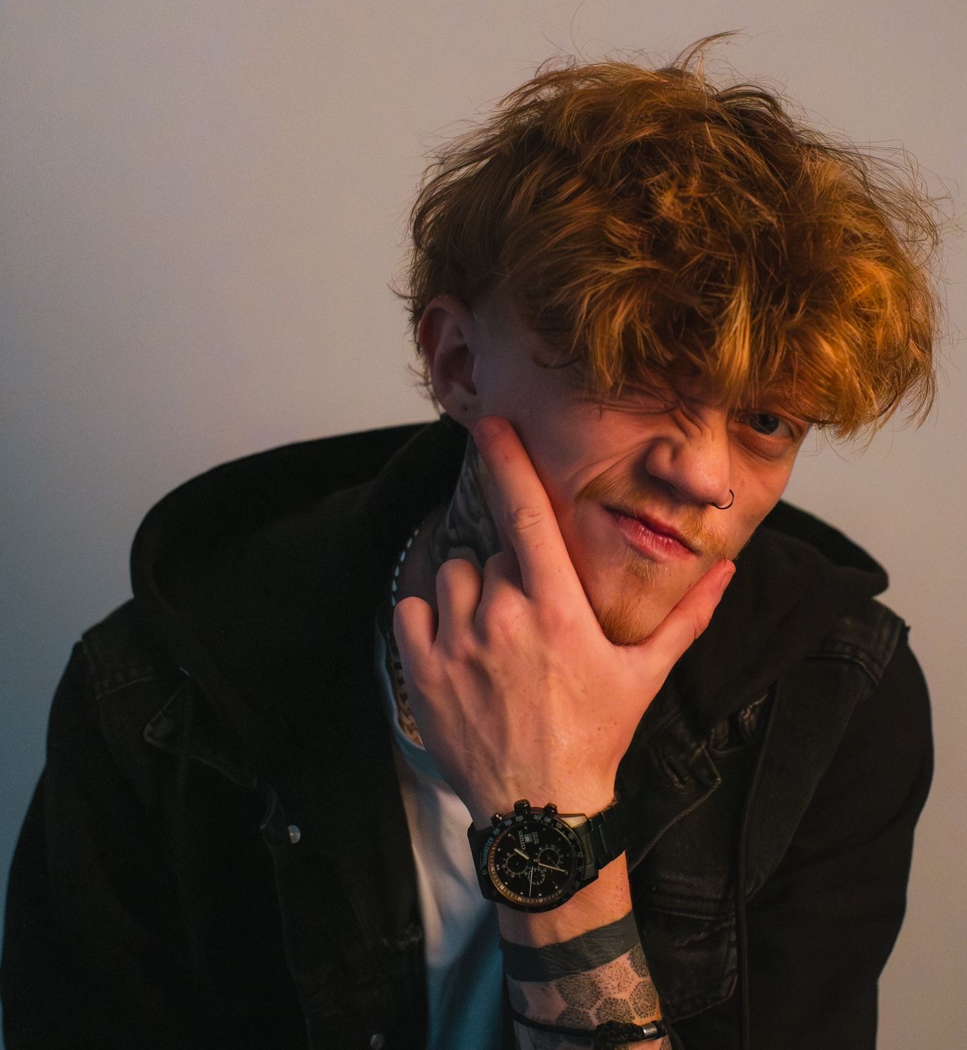 Young man with tousled light brown hair wearing a black jacket and watch, making a thinking gesture with his hand on his chin.