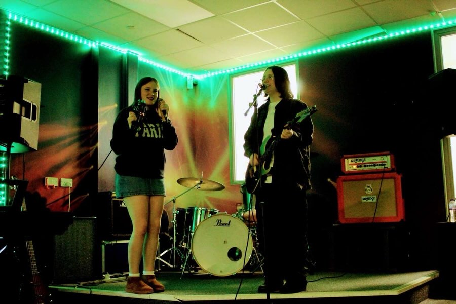 Two musicians on stage with drum set, one singing into microphone and tambourine, the other playing electric guitar singing with neon green ceiling lights.