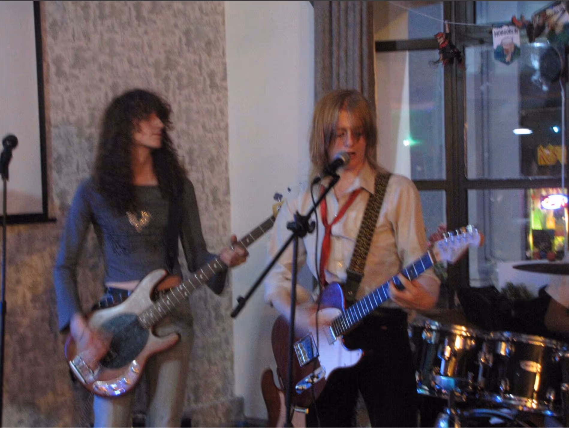 Two musicians performing indoors, one playing bass guitar and the other singing into a microphone while playing electric guitar.