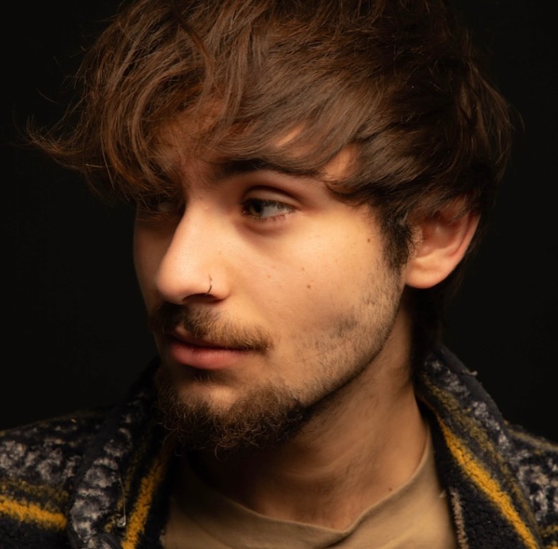 Young man with tousled brown hair, light facial hair, and a nose ring looking to the side against a dark background.