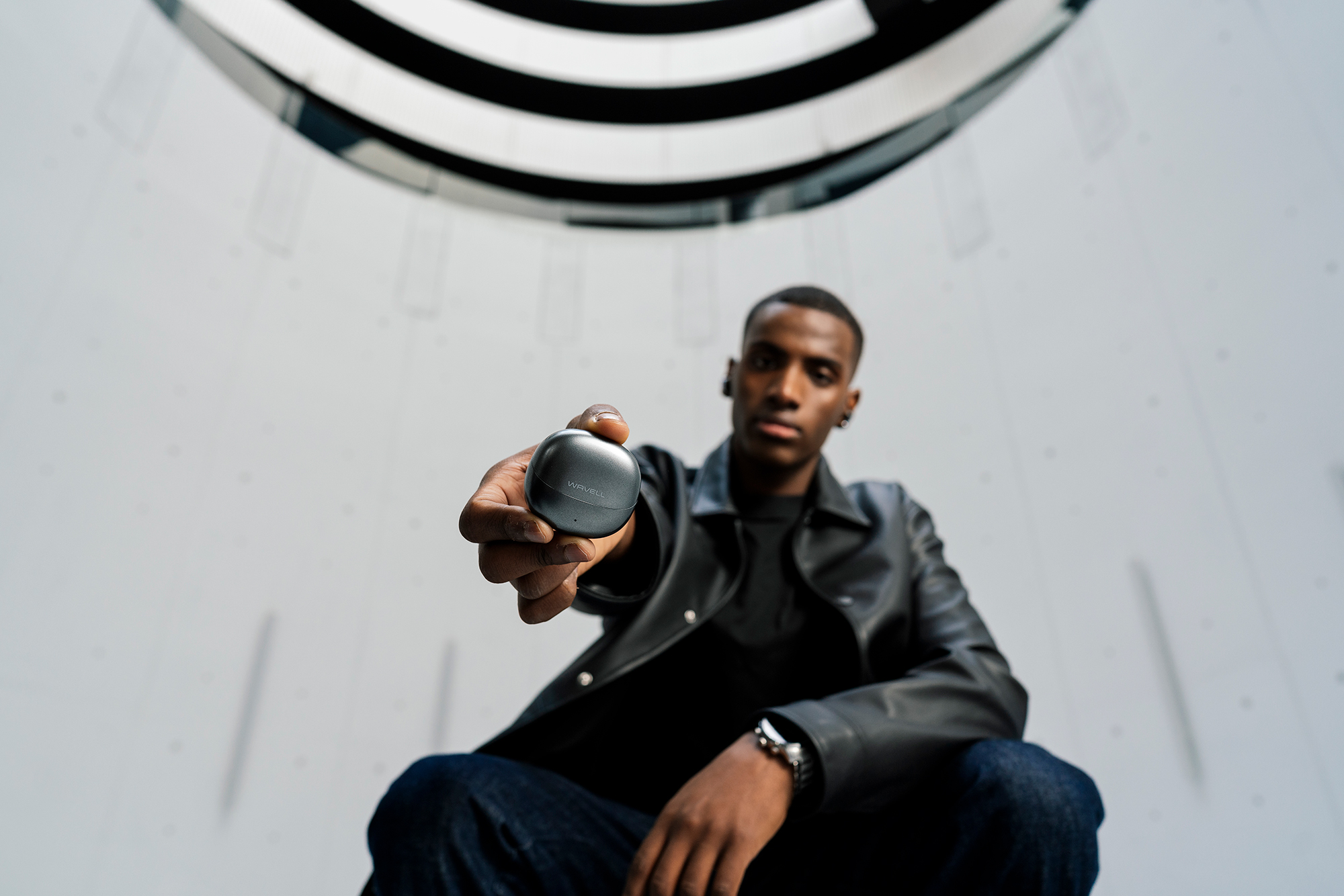 Man in black leather jacket holding a Wavell branded wireless earbud case toward the camera.