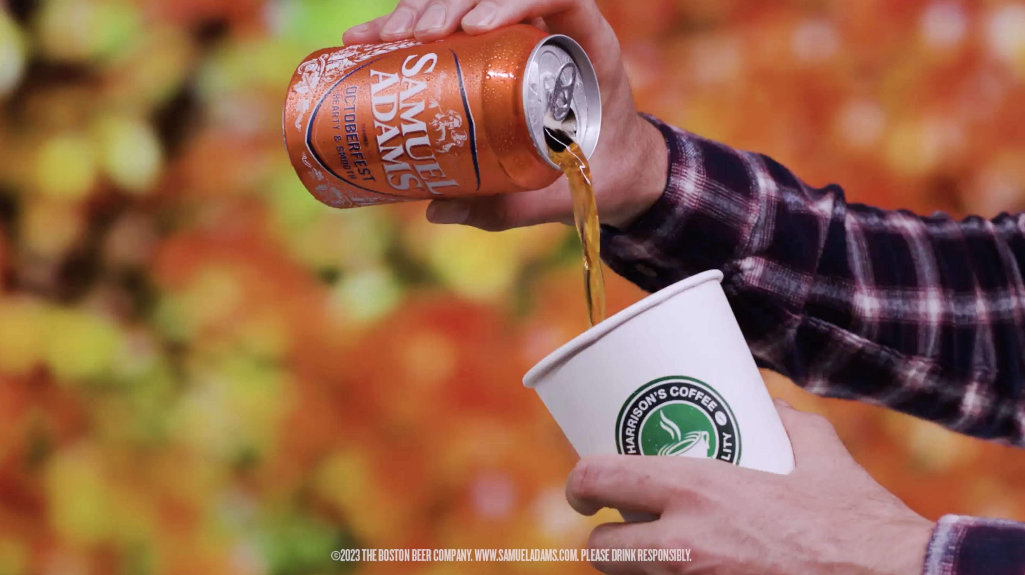Person in a plaid shirt pouring Samuel Adams Octoberfest beer from an orange can into a white Harrison's Coffee cup against a blurred fall foliage background.