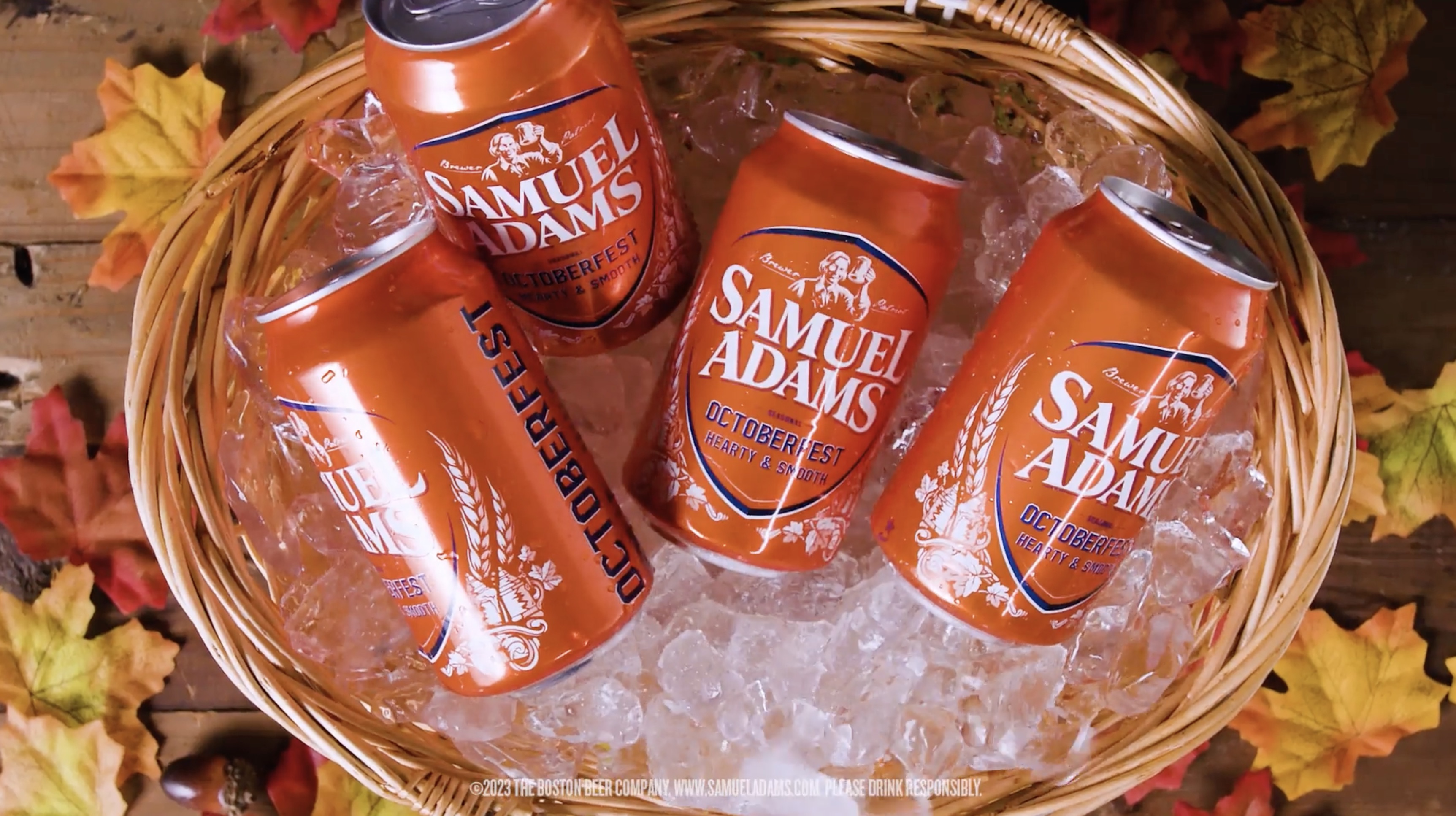 Four cans of Samuel Adams Octoberfest beer chilling in a basket filled with ice, surrounded by autumn leaves.
