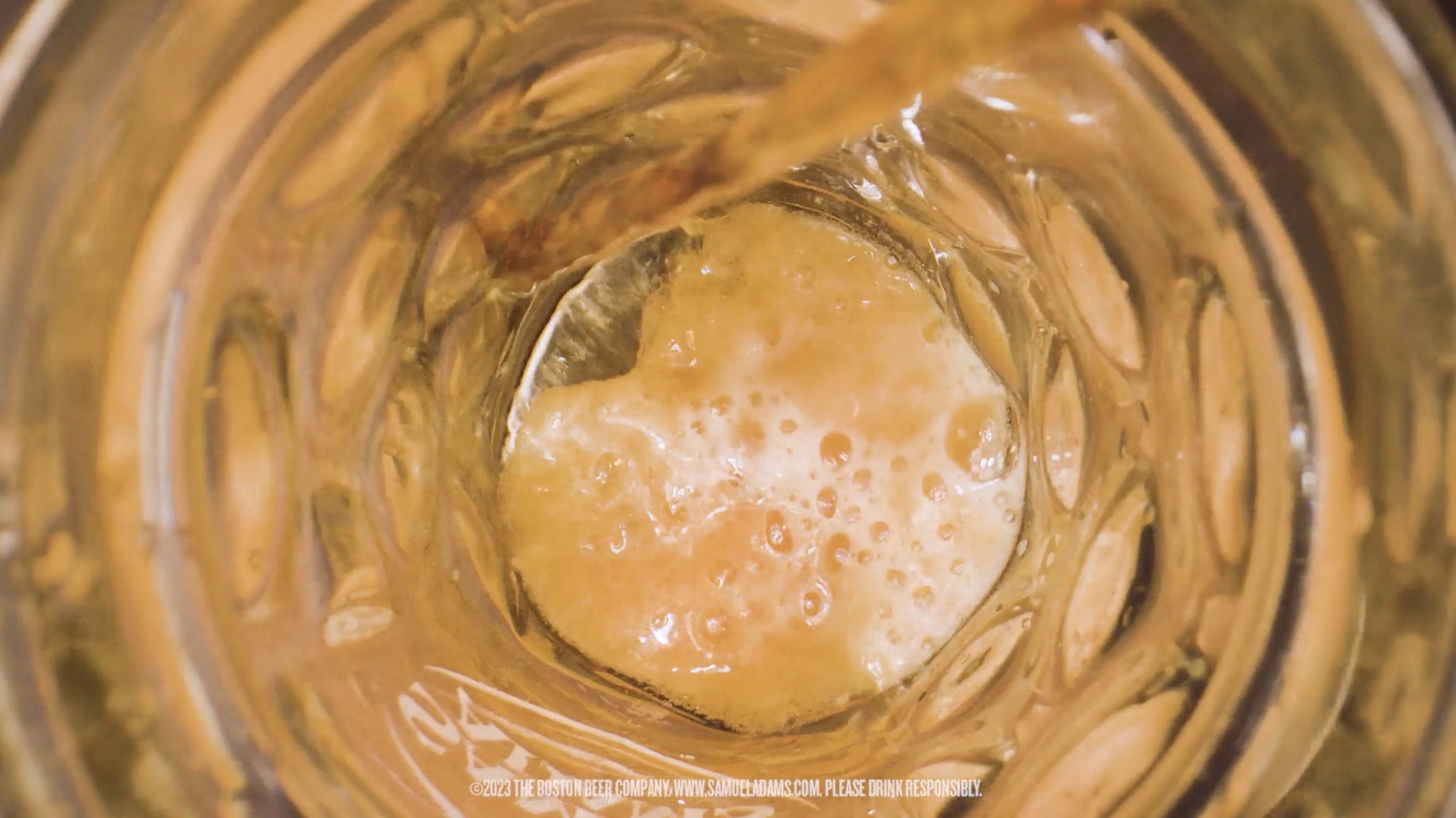 View into a glass mug showing amber beer being poured with foam forming at the bottom.