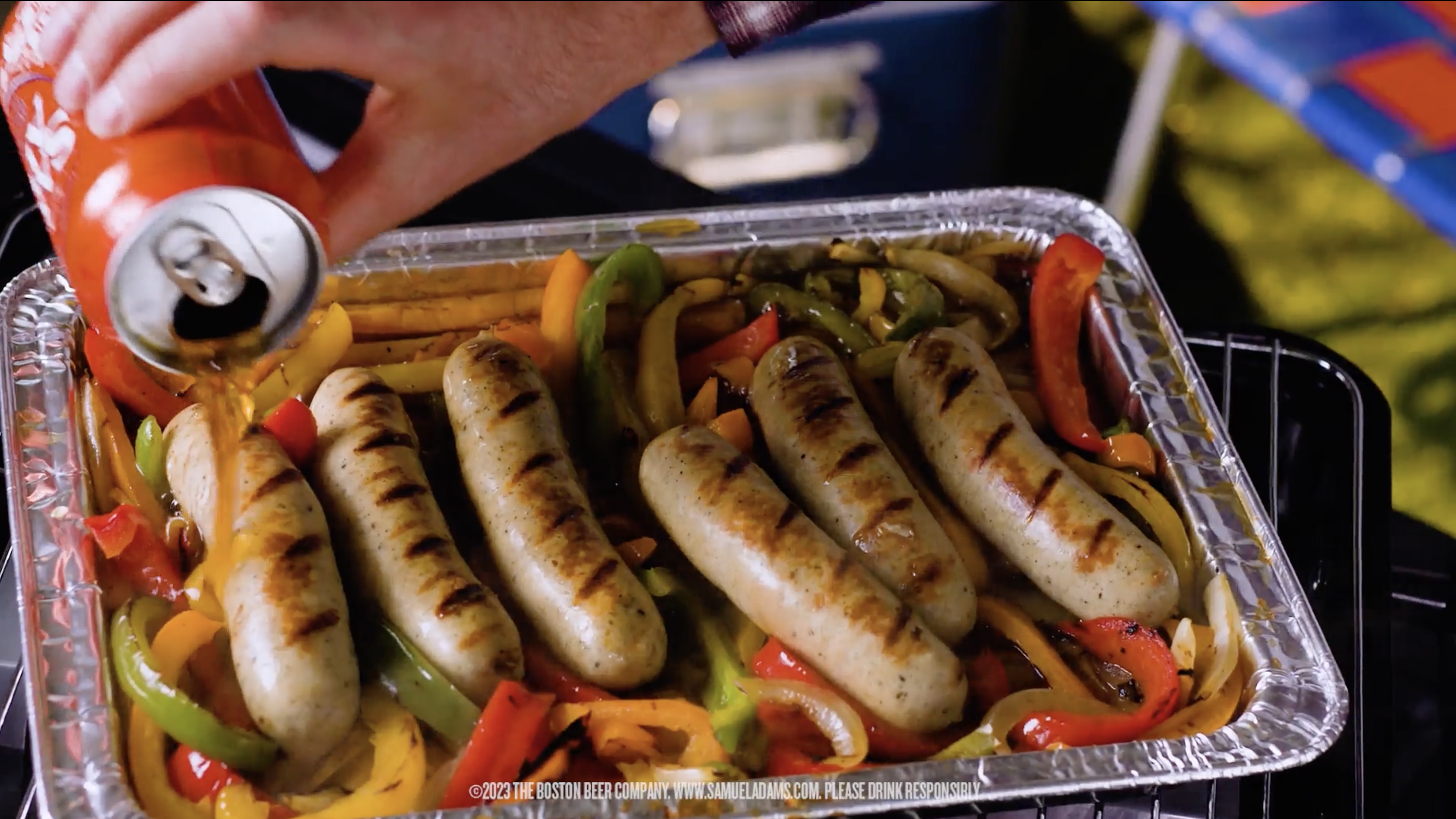 Grilled sausages and mixed bell peppers in an aluminum tray with a hand pouring liquid from an orange can.