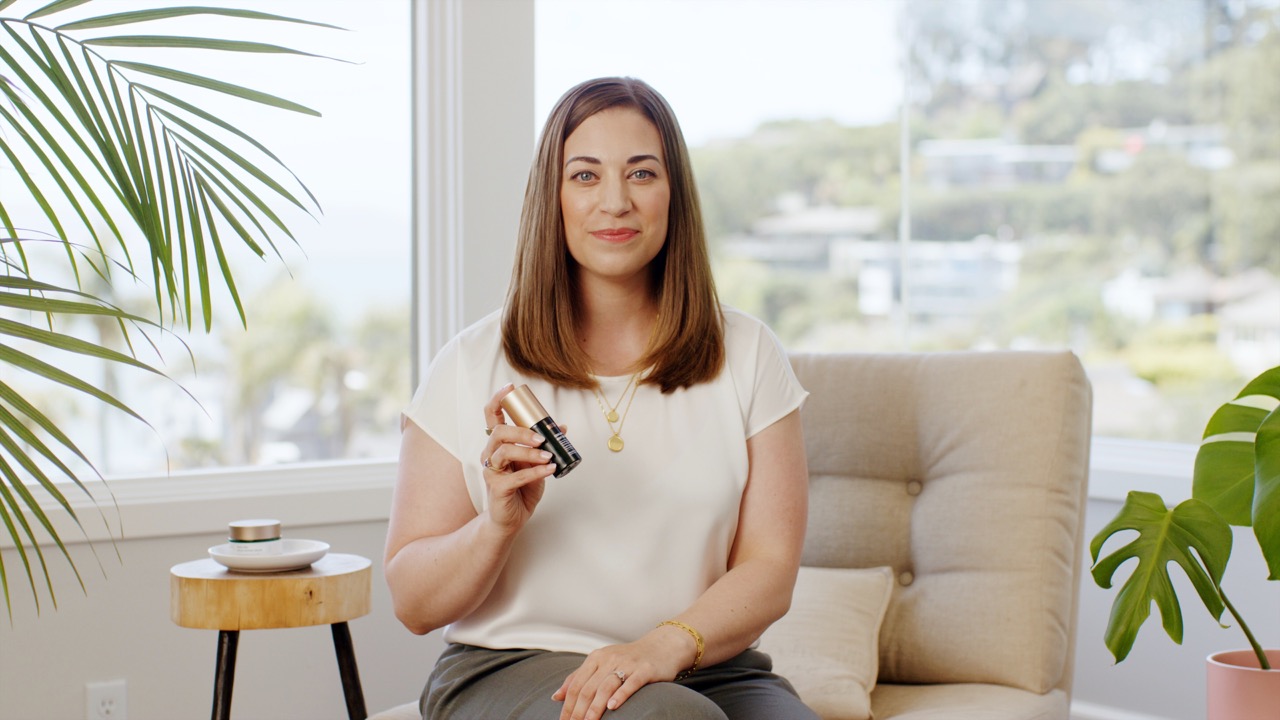 Smiling woman sitting on a beige chair in a bright room holding a dark bottle with a gold cap.