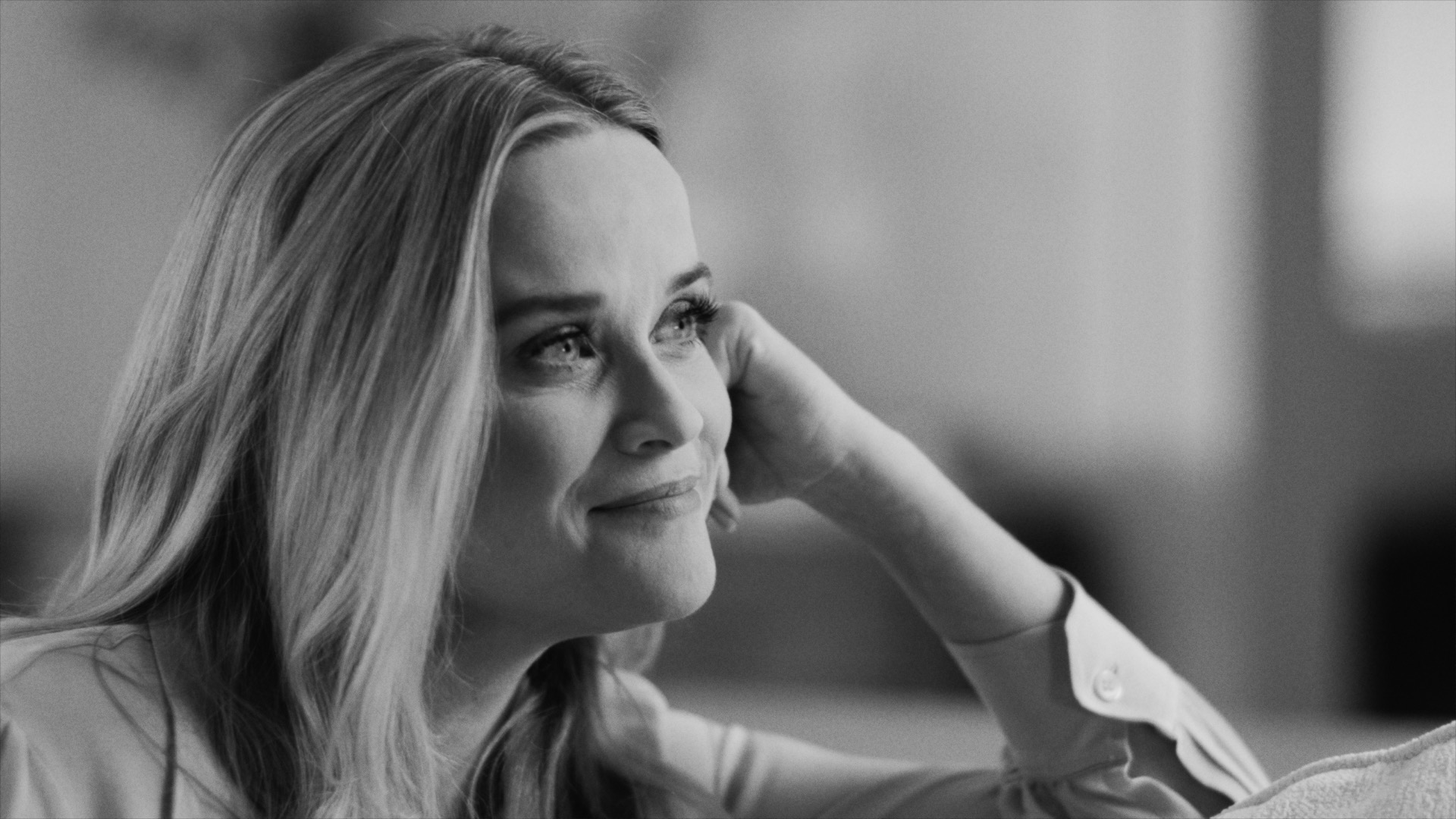 Black and white close-up of a Reese Witherspoon resting her head on her hand and smiling thoughtfully.