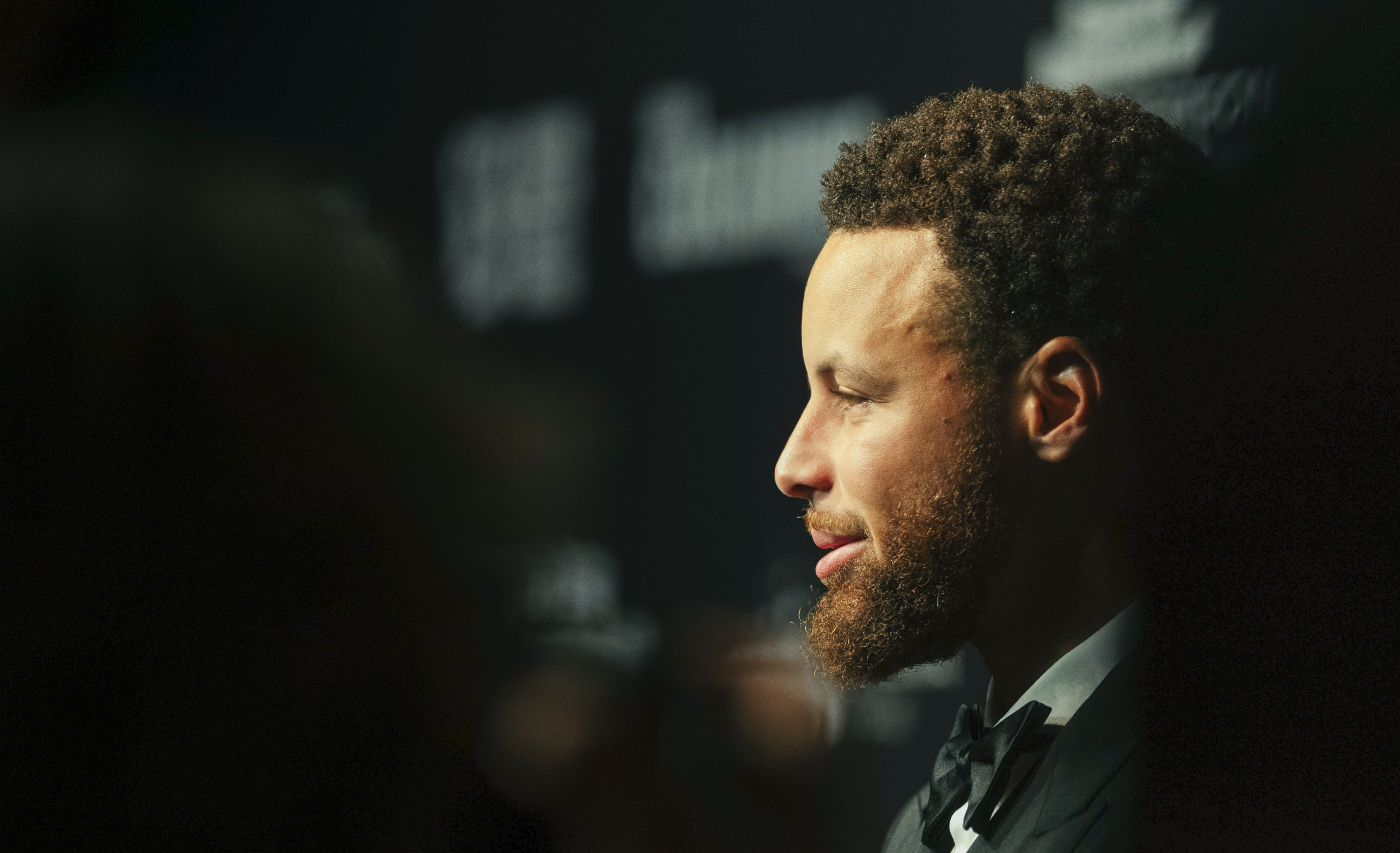 Side profile of Stephen Curry wearing a black tuxedo and bow tie against a dark background.