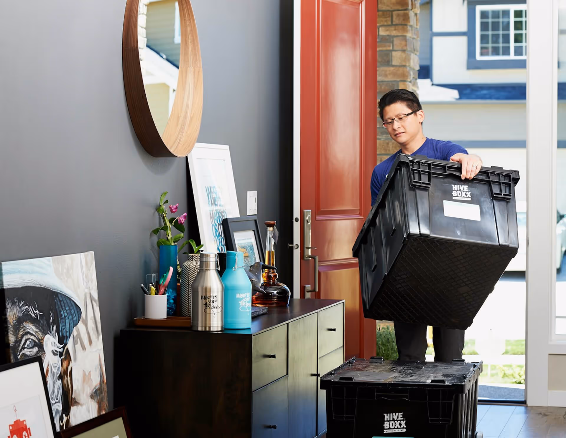 Man carrying a black Hive Boxx container into a house with a red door and a decorated console table inside.