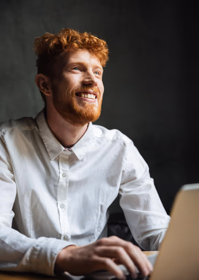 Smiling red-haired man in white shirt working on laptop against dark background.
