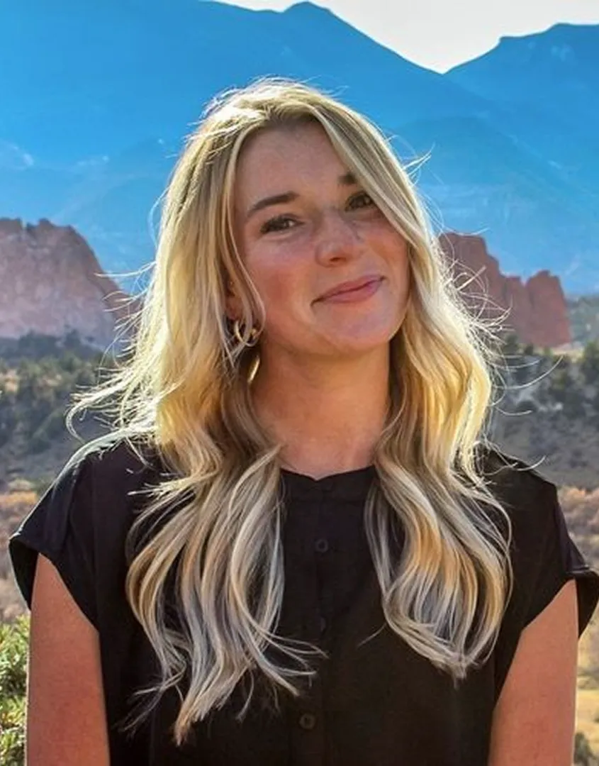 Smiling woman with long blonde hair wearing a black shirt outdoors with mountains in the background.