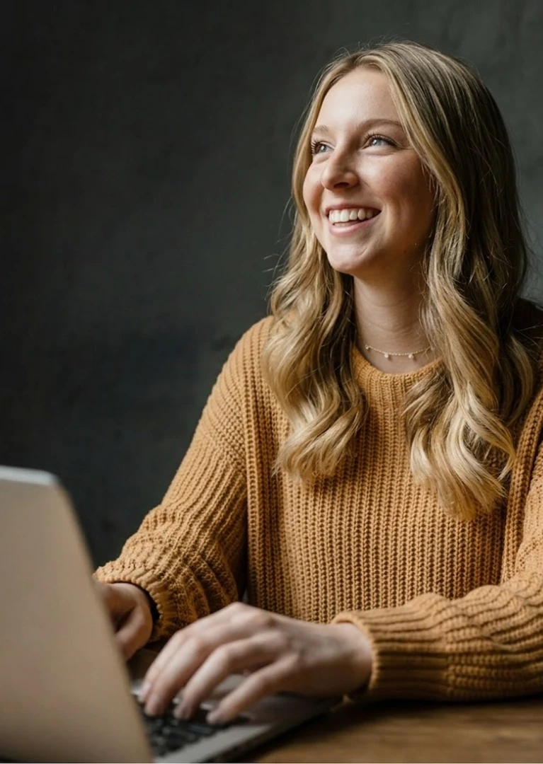 Smiling woman with blonde hair in a mustard sweater typing on a laptop at a wooden table.