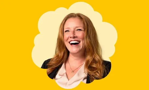 Smiling woman with long red hair wearing a black jacket and white blouse against yellow background.