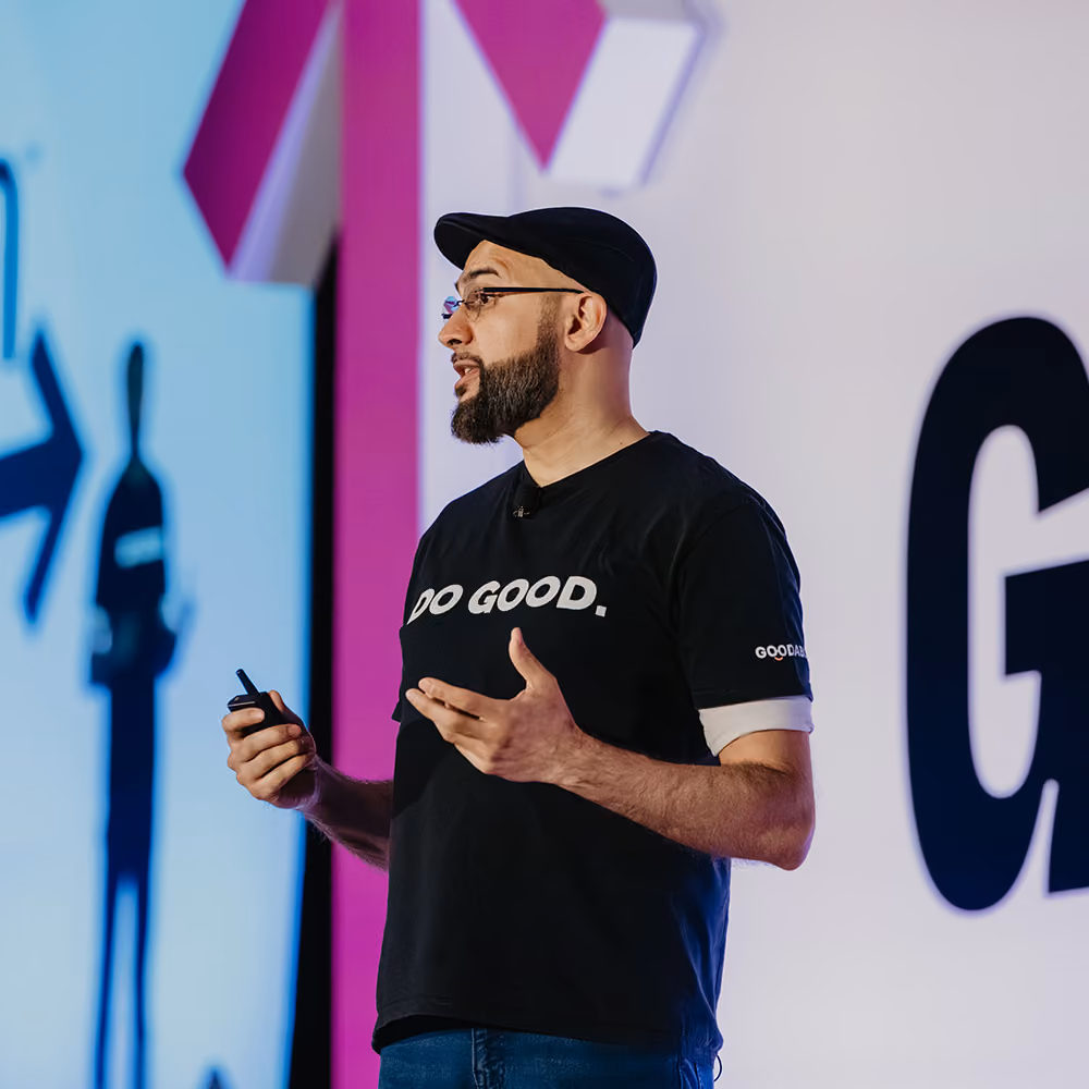 Man wearing a black cap and Do Good t-shirt speaking on stage with a microphone and presentation screen at GiveCon