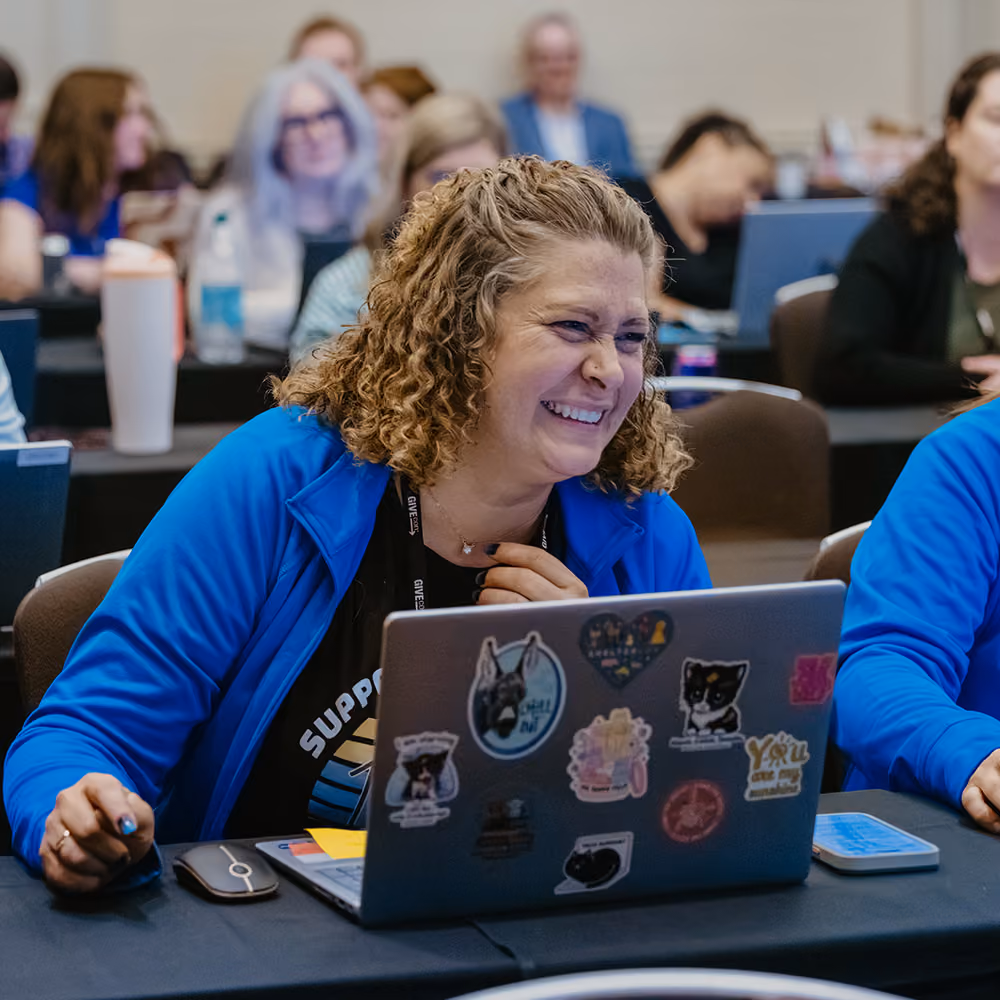 Smiling woman in blue jacket working on a laptop covered with stickers at GiveCon