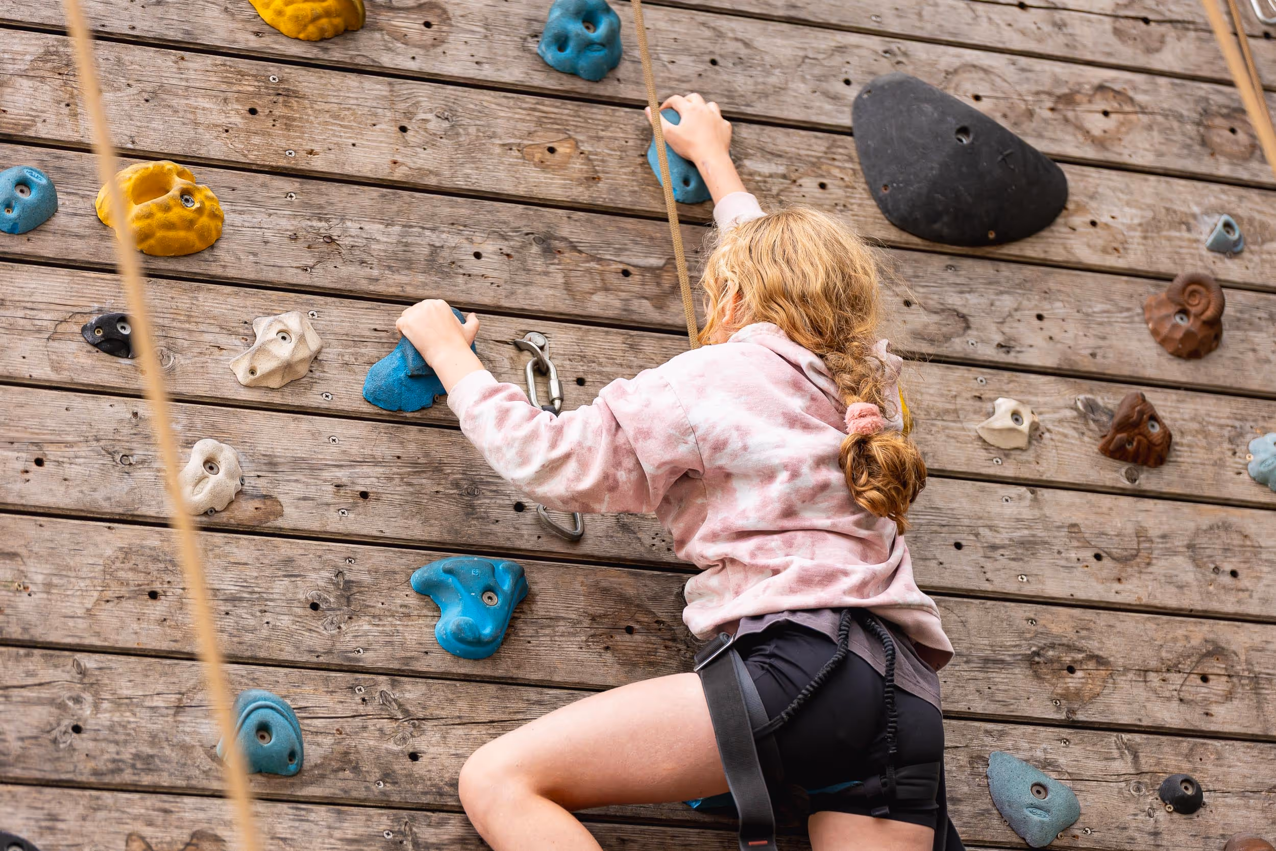 Child climbing a rock climbing wall, wearing a pink hoodie and safety harness.