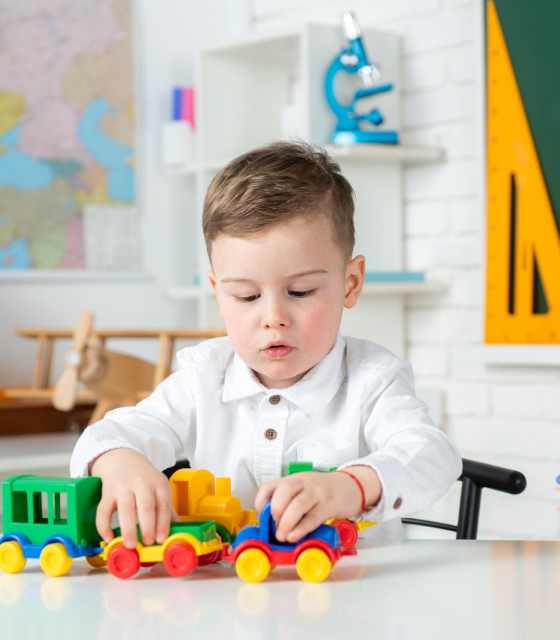 Young boy in white shirt playing with colorful toy train at a table in a classroom.